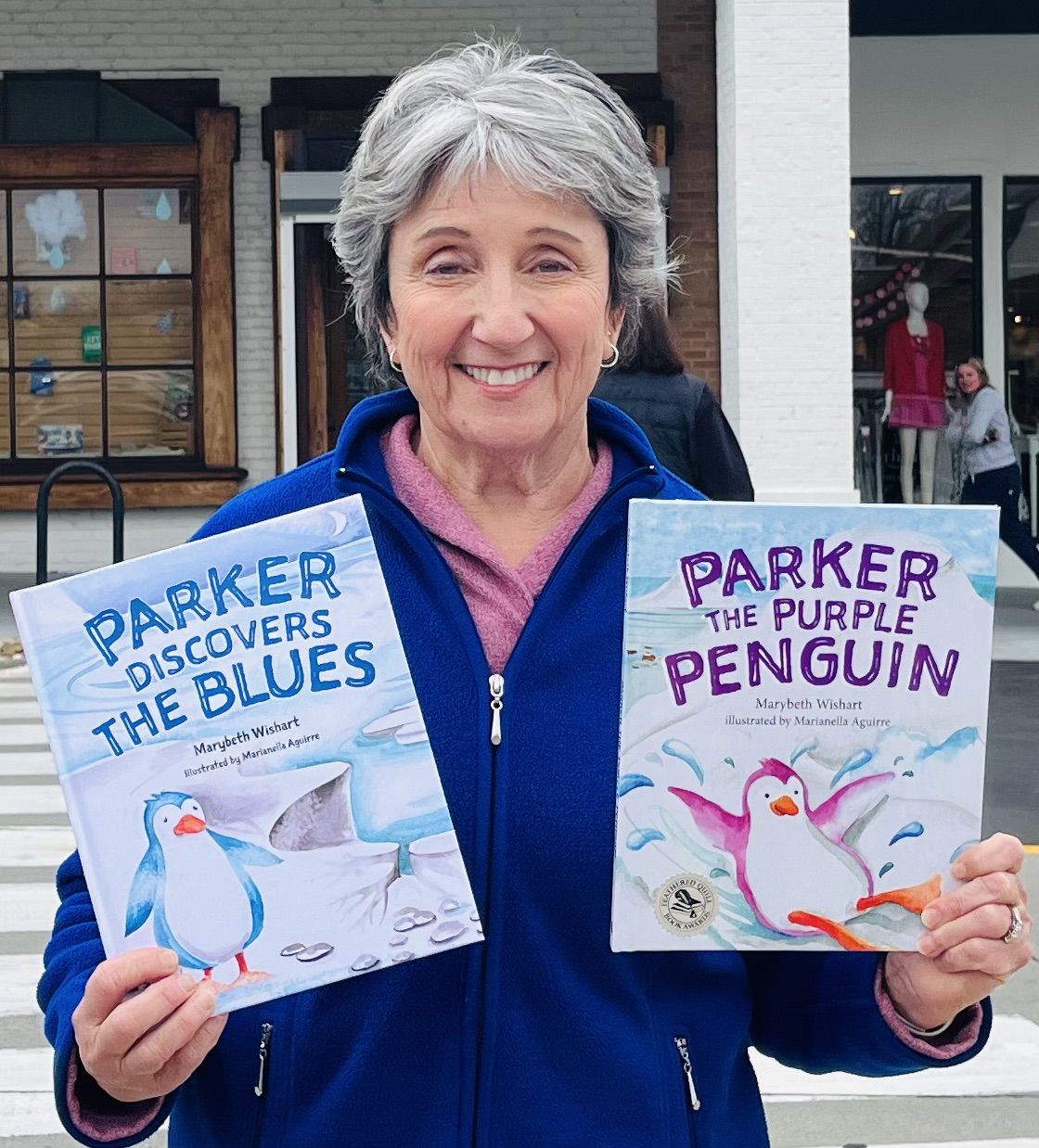 Smiling woman holding two children's books about penguins outdoors, storefronts in background.