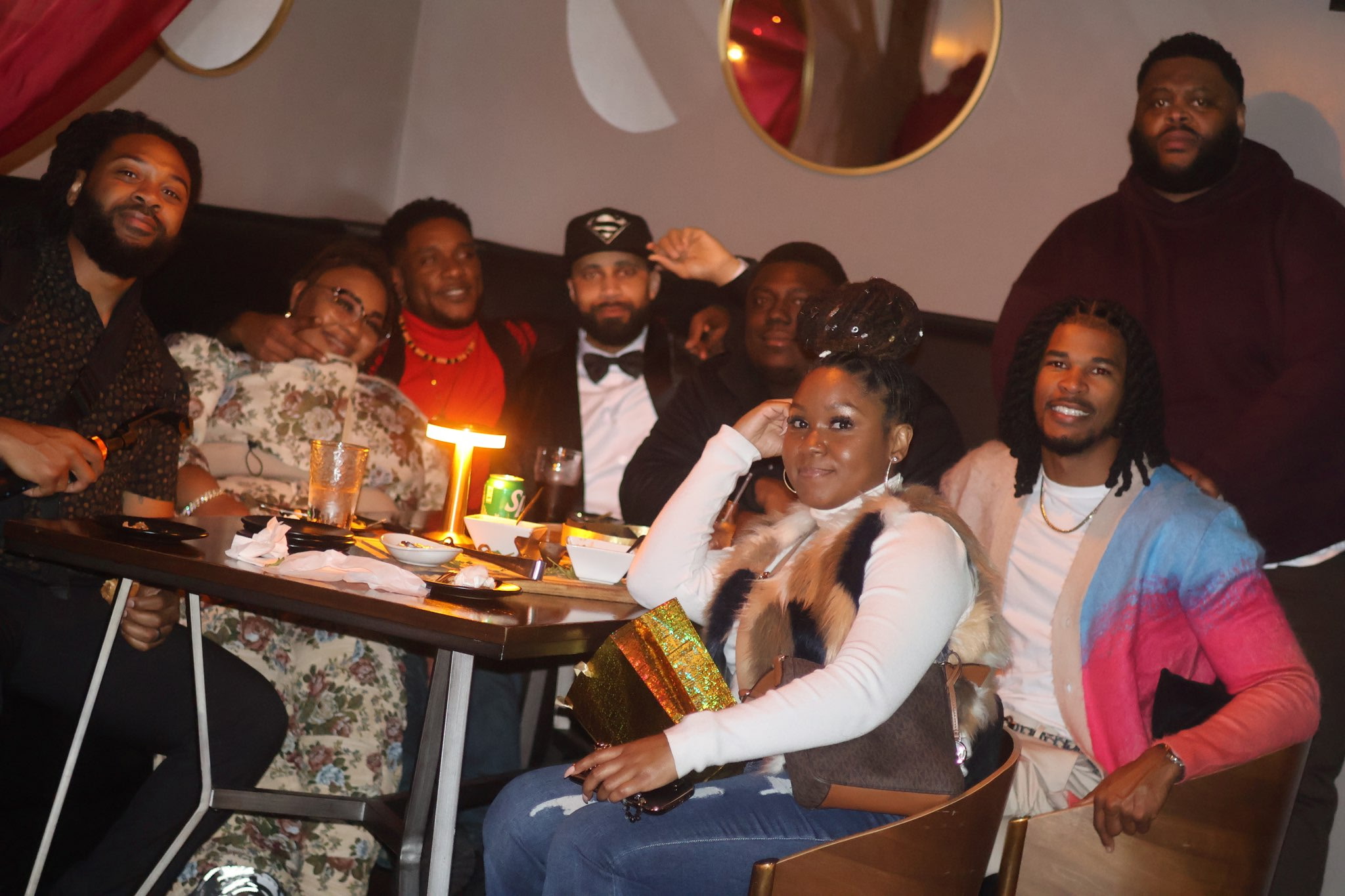 Group of eight people celebrating at a table with a birthday cake and lit candle, smiling and posing for the photo.