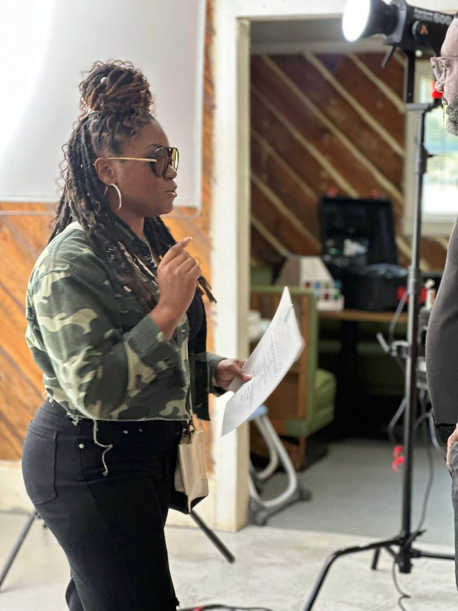 Woman with dreadlocks and sunglasses holding a paper, standing in a room with studio equipment and wooden wall.