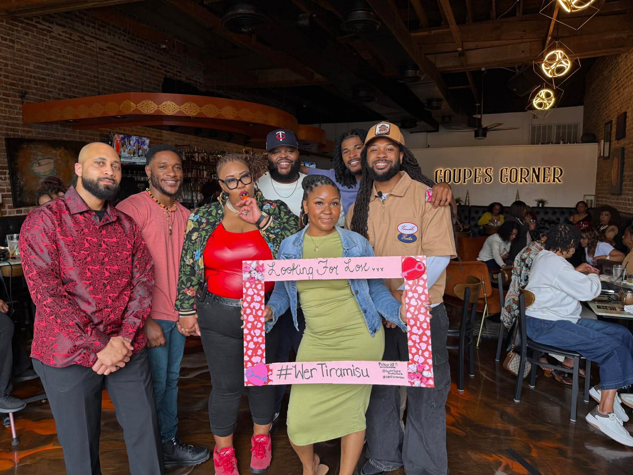 Group of seven diverse people standing inside a restaurant, holding a pink frame with handwritten text, smiling at the camera.