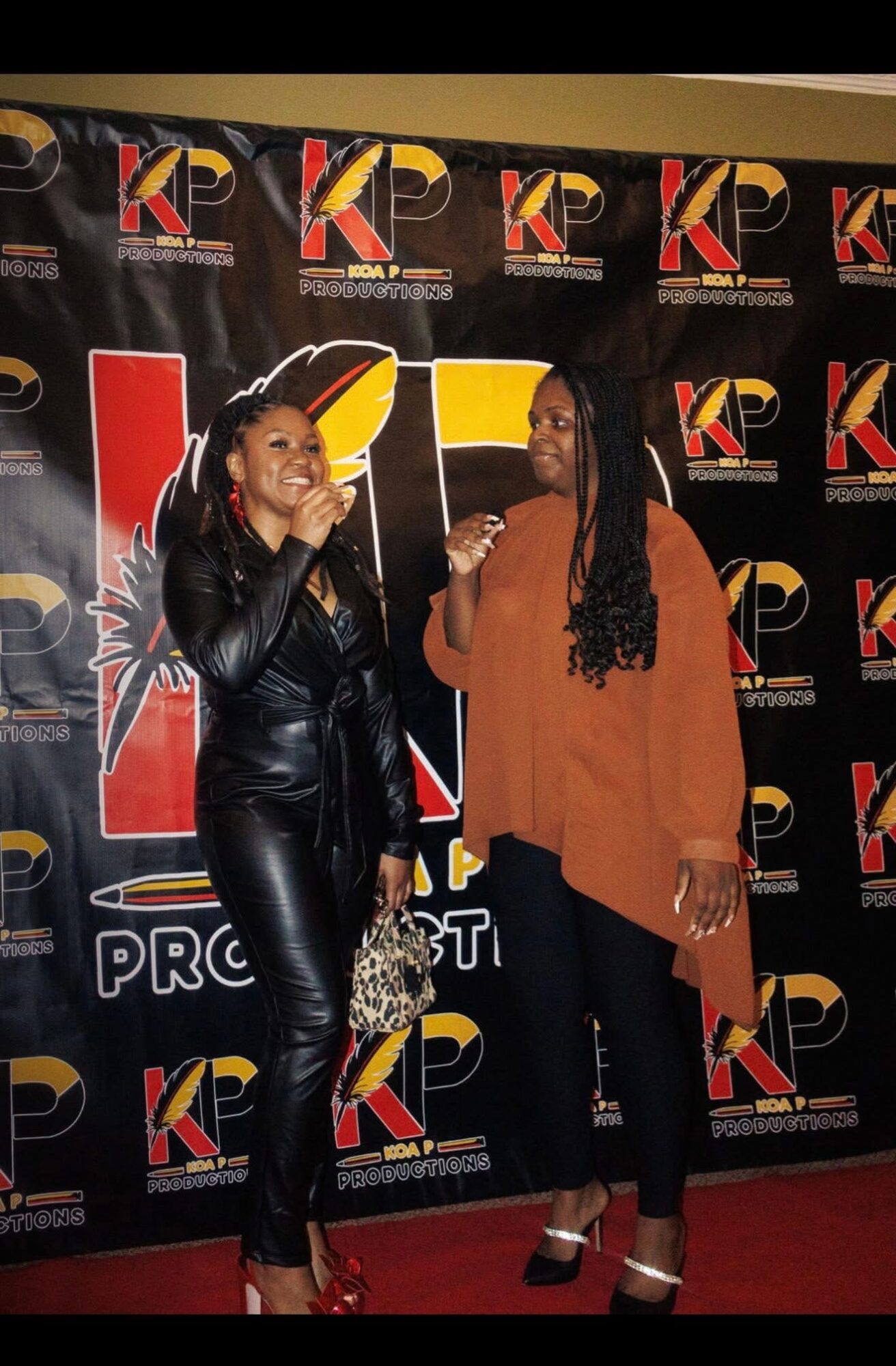 Two women stand in front of a black backdrop with colorful logos, smiling and talking to each other.