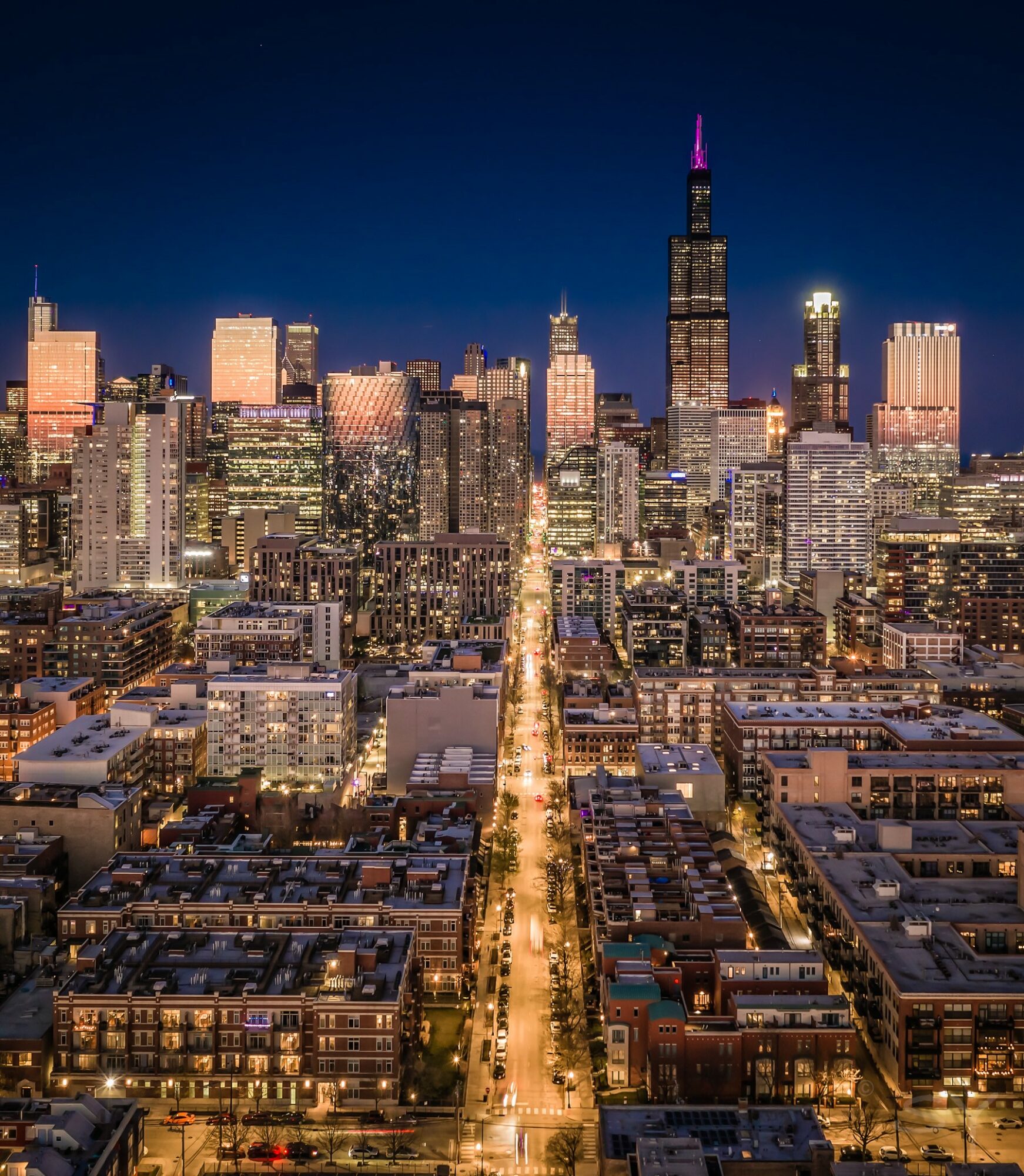 Nighttime city skyline with illuminated buildings and a central street leading to a tall skyscraper with a purple-lit spire.