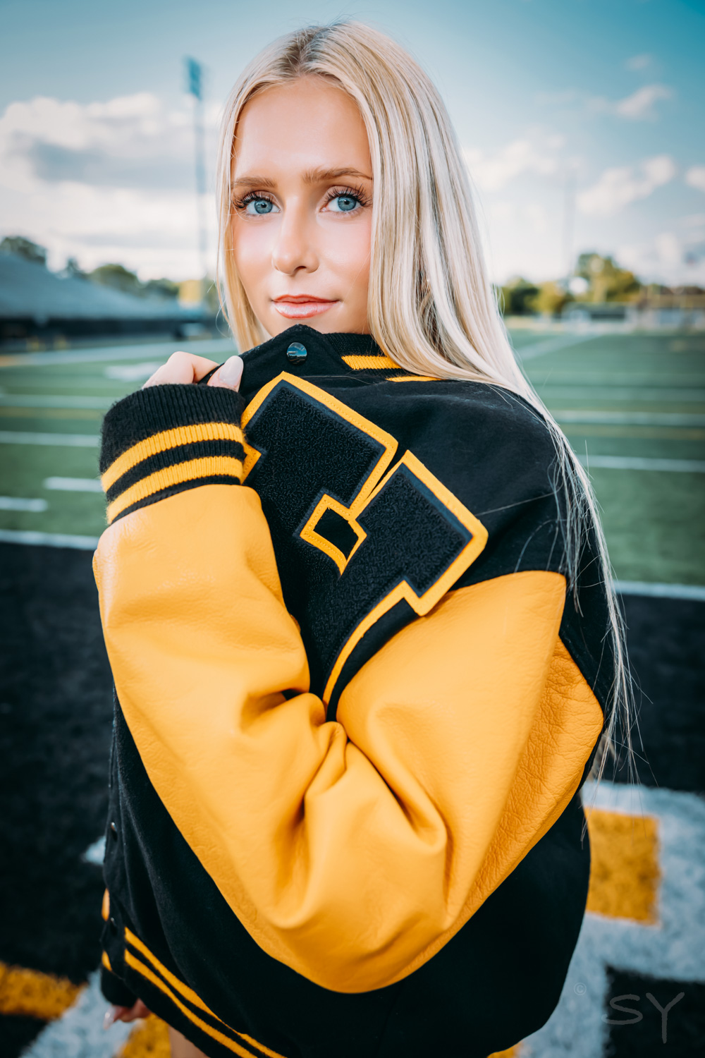 Young woman with long blonde hair wearing a black and yellow sports jacket, standing outdoors on a field.