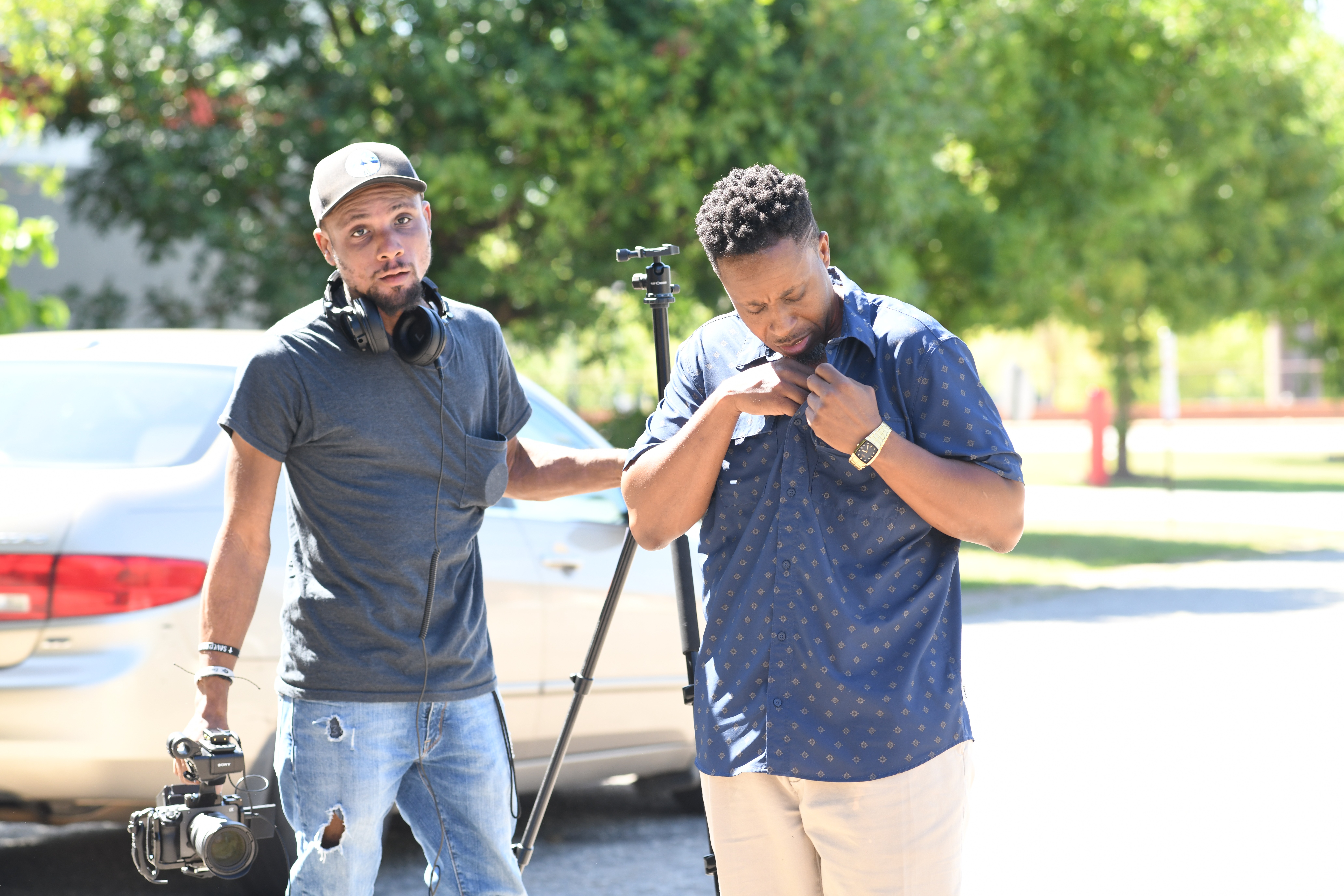Two young men standing outdoors near a car, one with headphones around his neck and the other looking at his phone, with trees in the background.