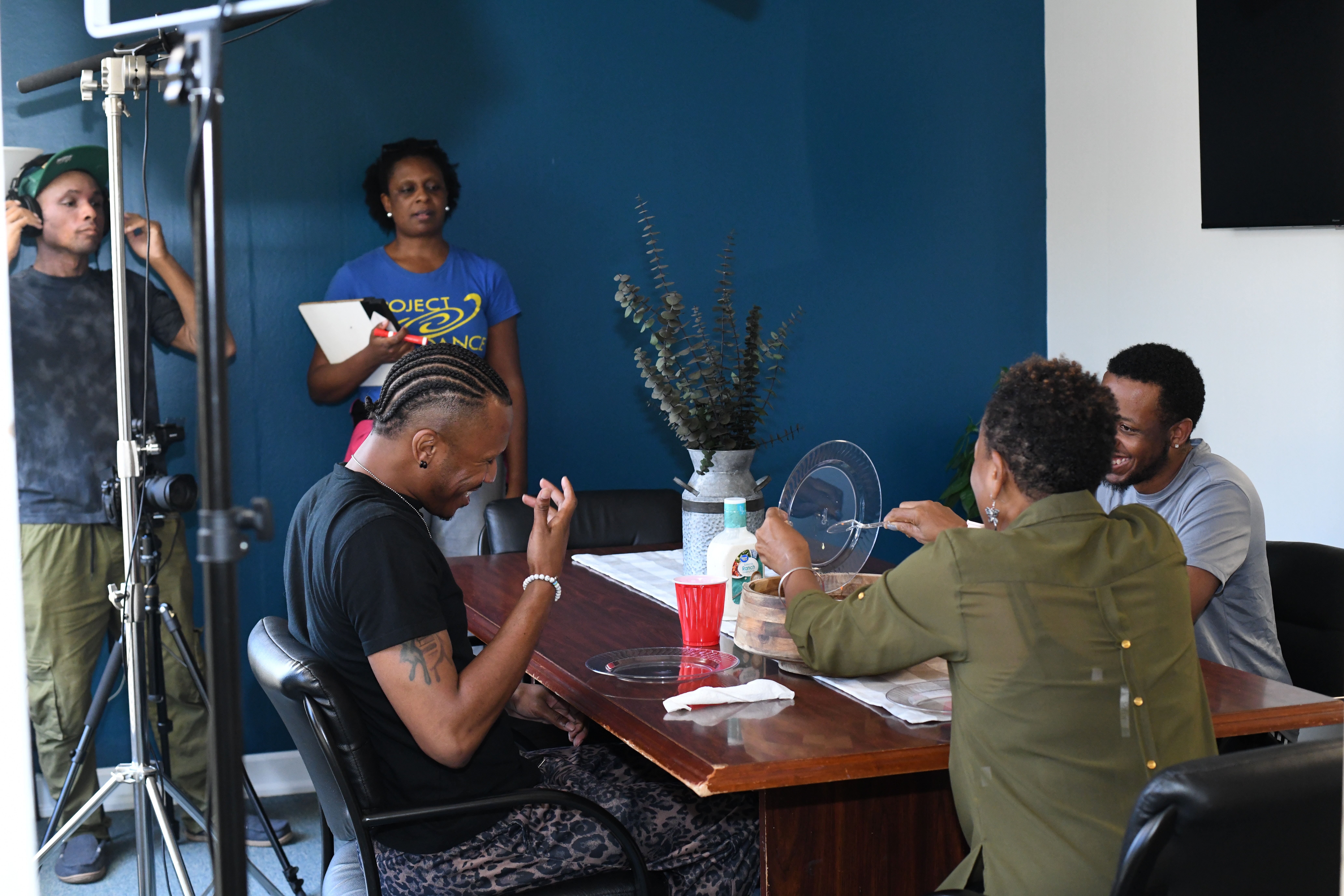 Four people are in a room with a blue wall, sitting around a table, with one person standing holding a notebook.