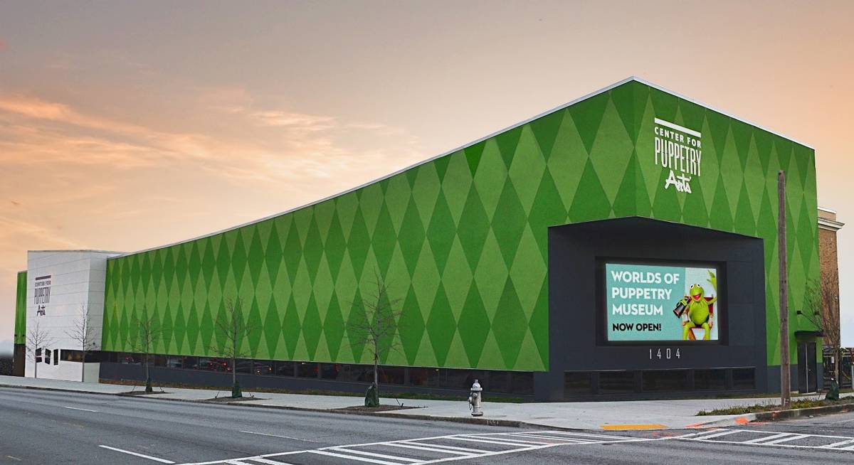 Large green building with a digital display and sign for a puppet museum, on a street with a sidewalk and road.