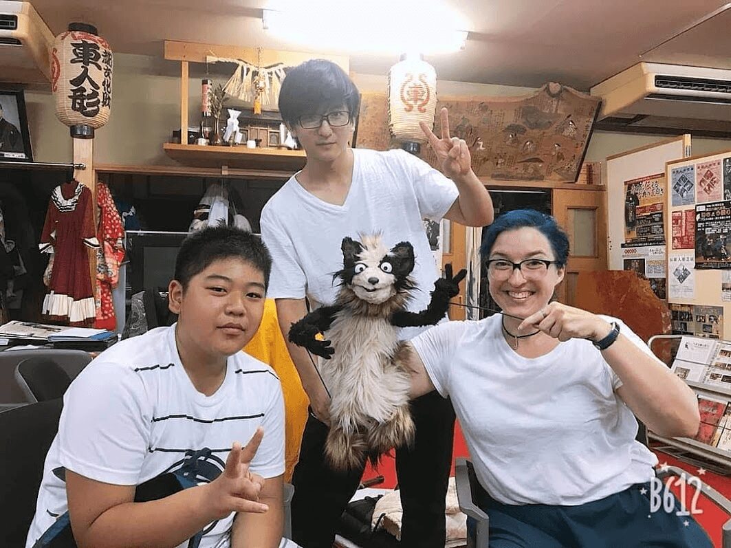 Three people and a dog in a room with decorations, one person making a peace sign, another holding the dog, smiling.