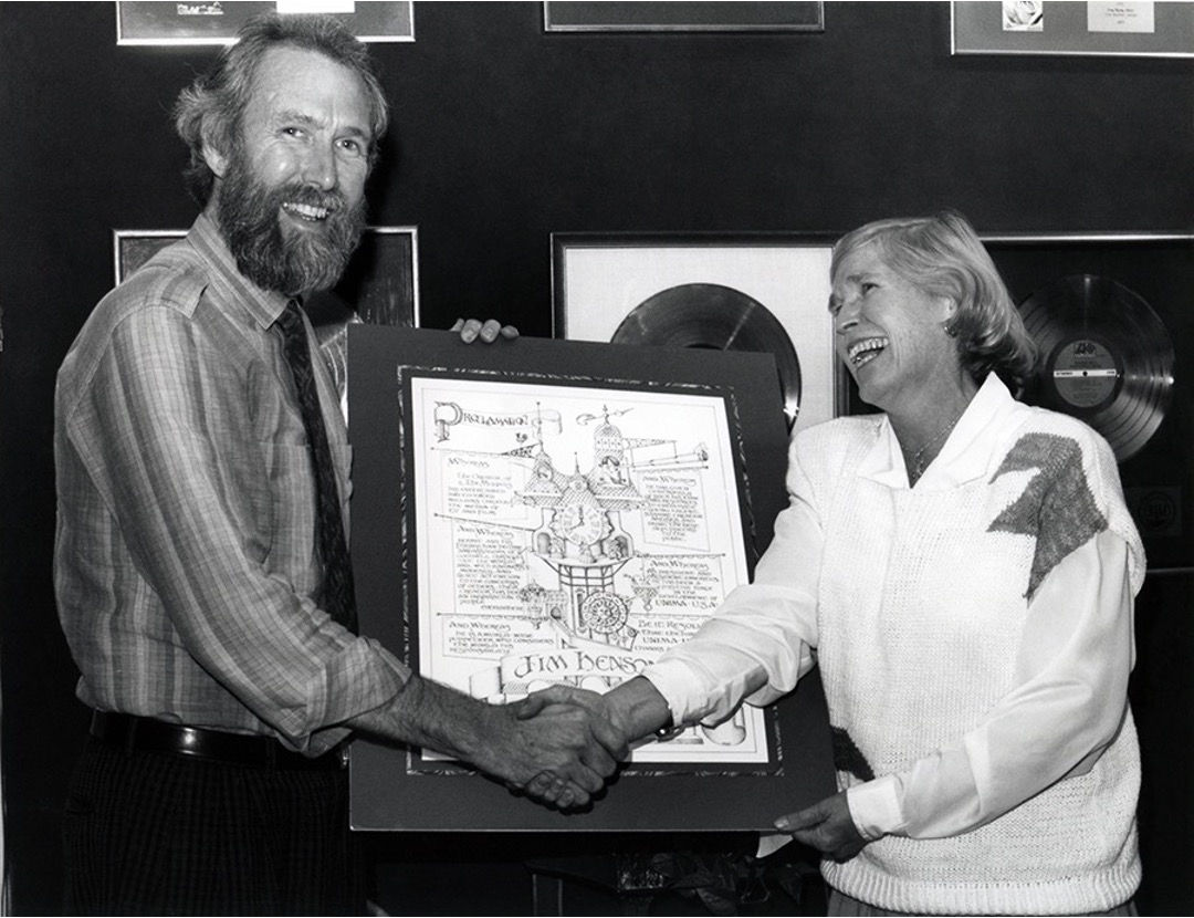 Two people shake hands, one holding a framed document, smiling in a room with framed records on the wall.