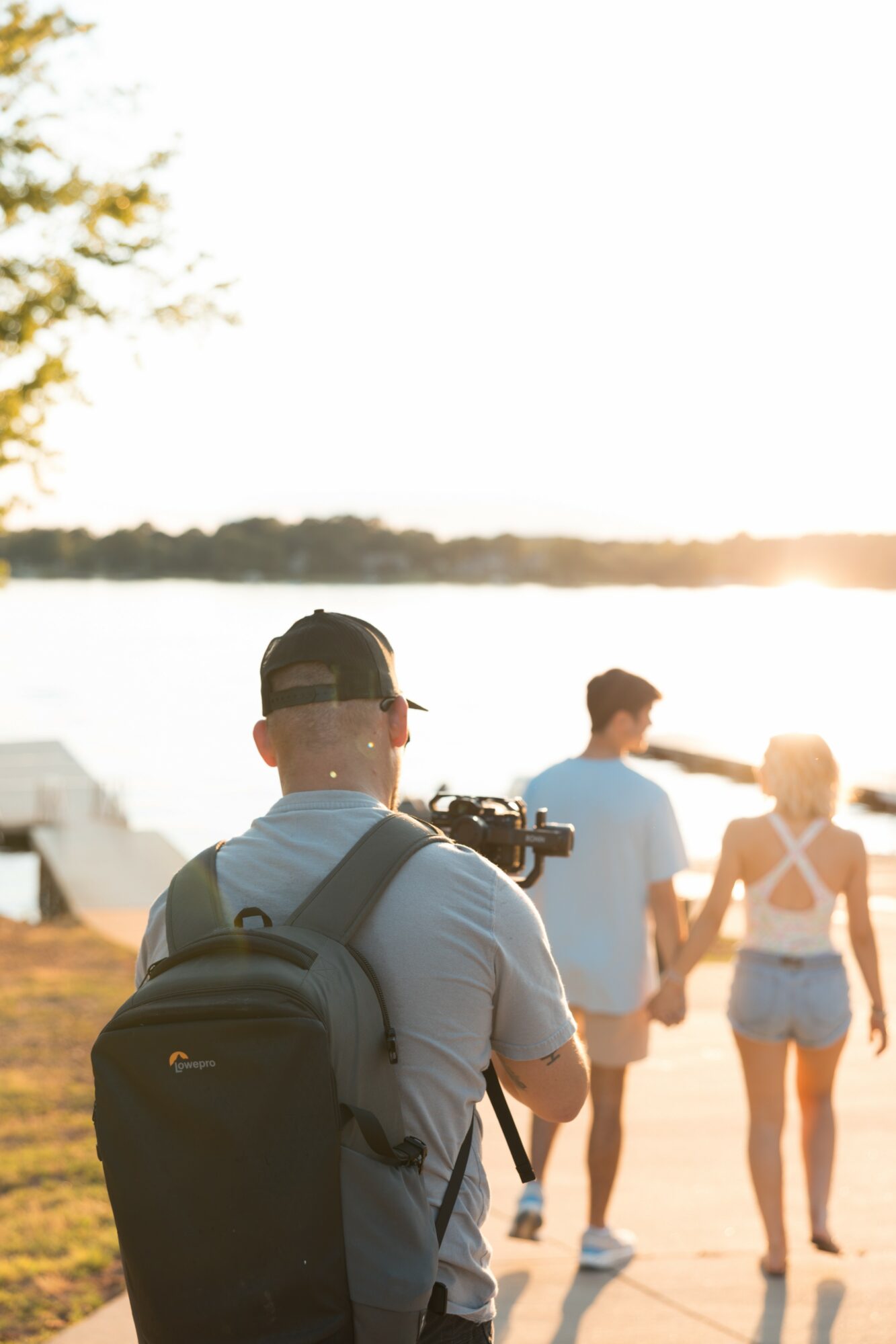 Group of people walking near a body of water during sunset, with a person in the foreground wearing a backpack and cap.