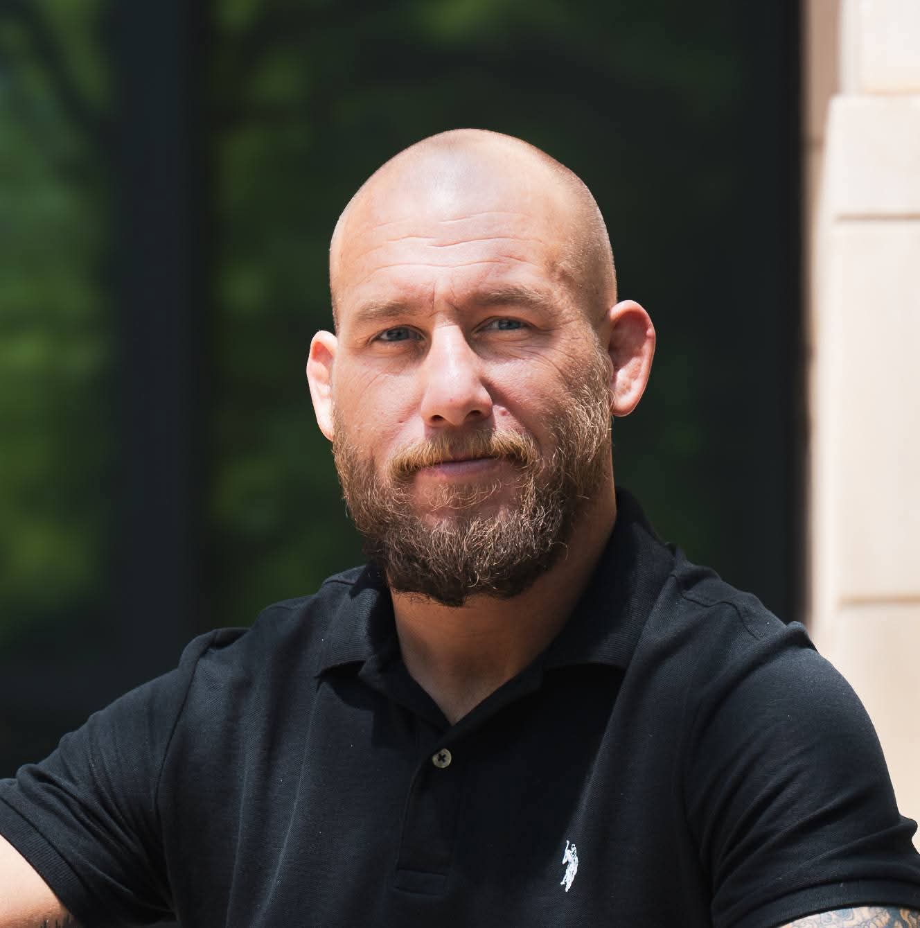 Bald man with beard wearing a black polo shirt outdoors, looking at the camera.