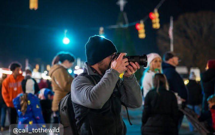 Person wearing a beanie and gray jacket taking a photo with a camera at night, surrounded by people and colorful lights.