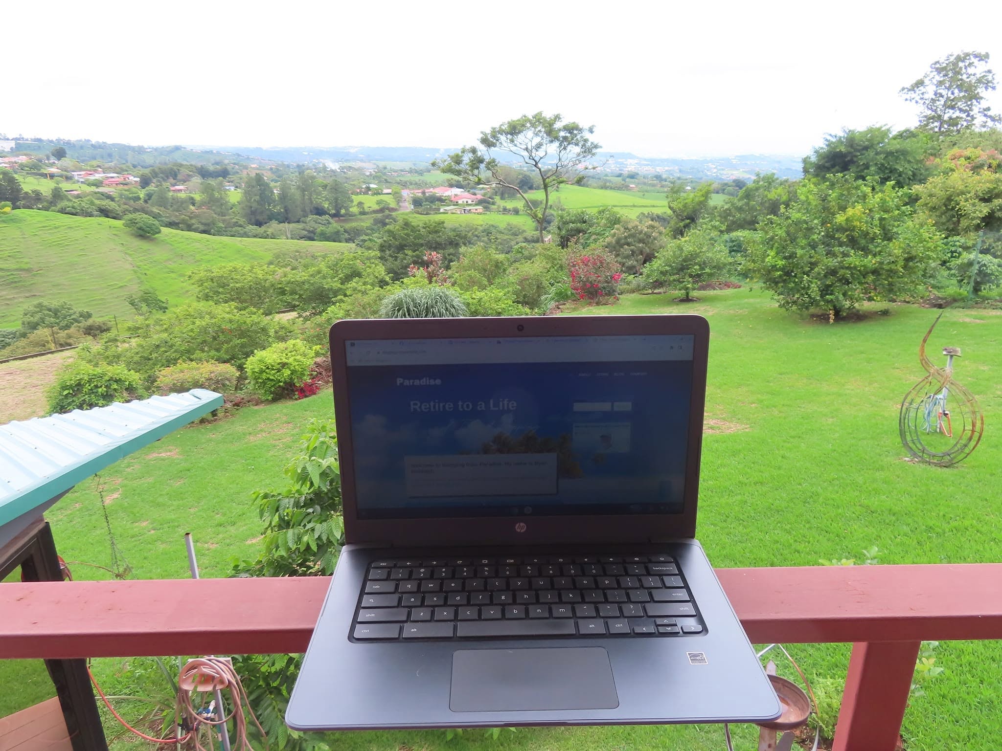 Laptop on a balcony railing overlooking a green landscape with trees and fields.
