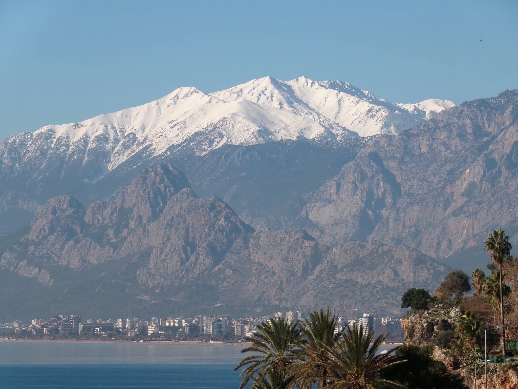 Snow-capped mountain range with lower rocky mountains and a cityscape along a body of water, trees in foreground.