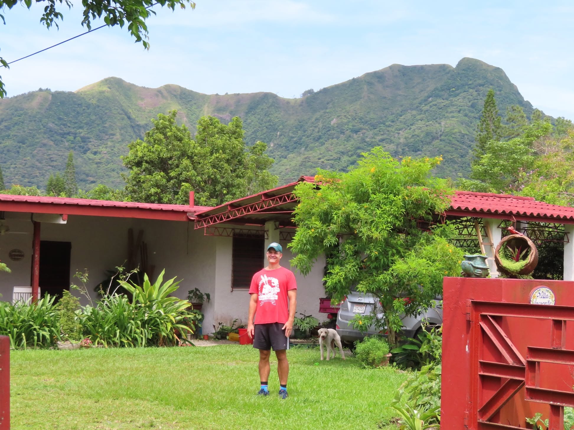 A person wearing a red shirt, black shorts, and a cap stands on a grassy yard with trees and mountains in the background.
