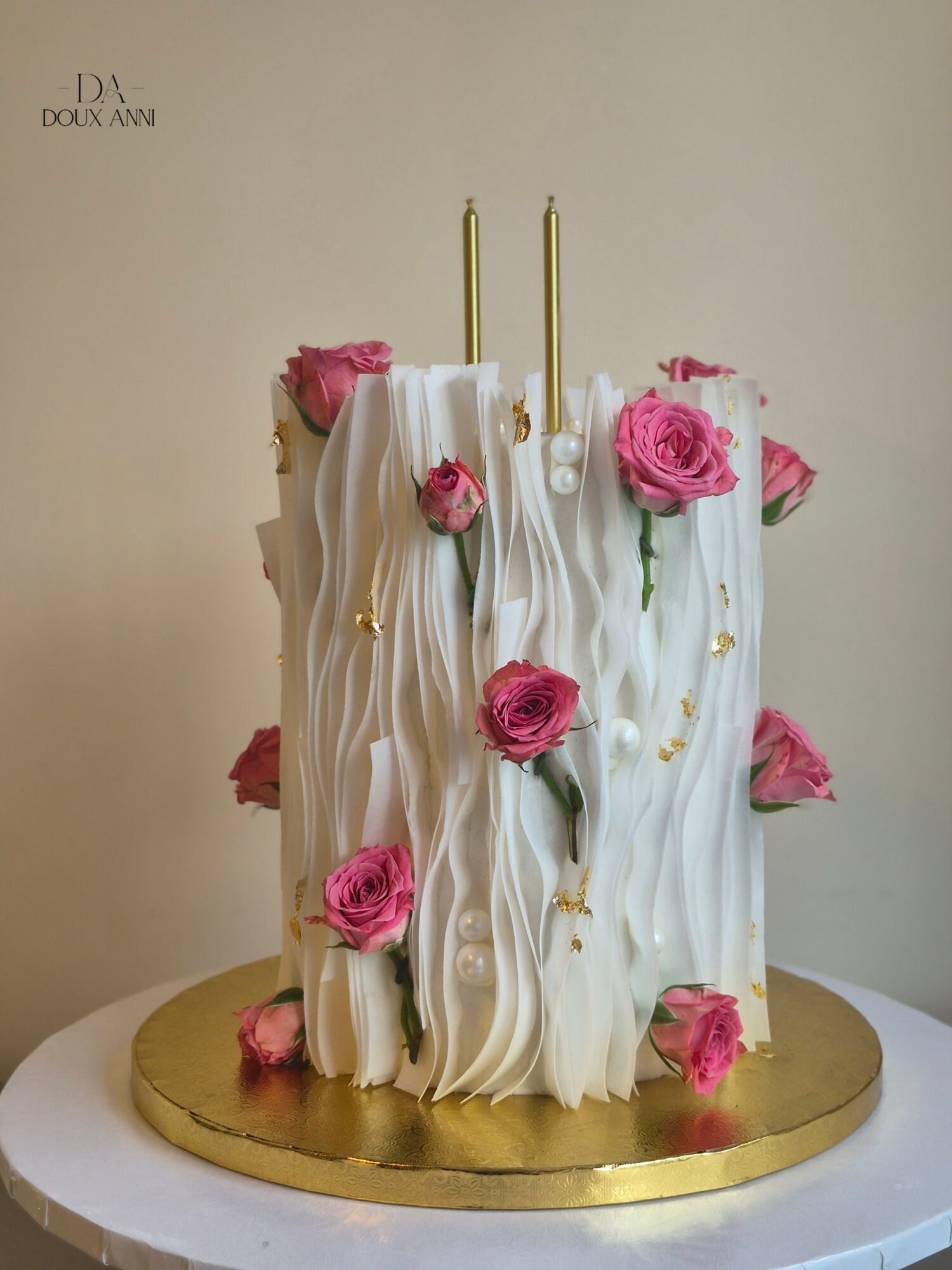 Tall white cake with pink roses and gold accents on a gold base, two gold candles on top.