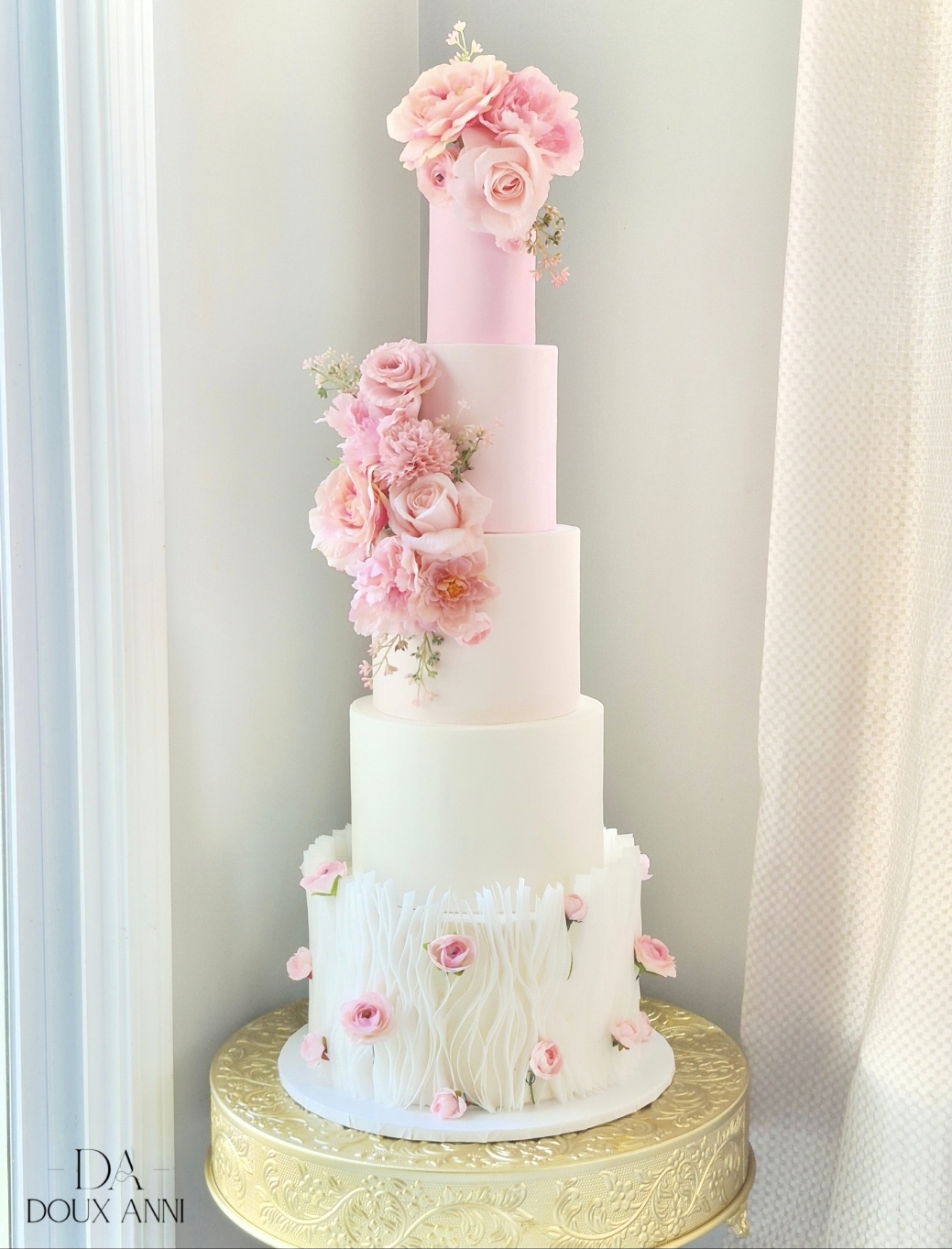 Tall white tiered cake decorated with pink flowers, placed on a gold stand, against a light background.