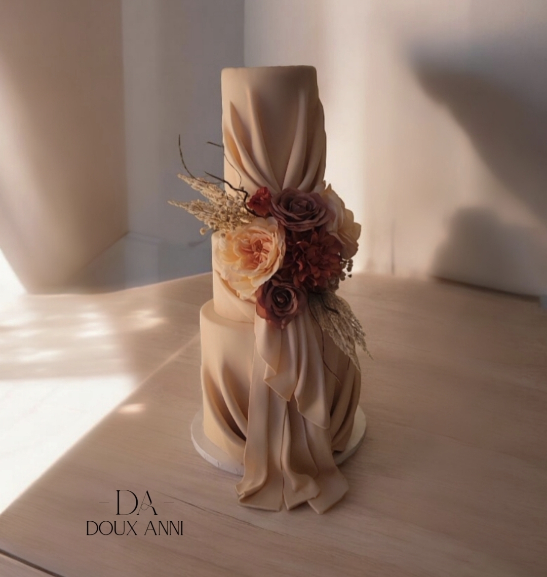 A tall, beige, fabric-covered candle with a floral arrangement and draped fabric, placed on a light-colored surface with sunlight.