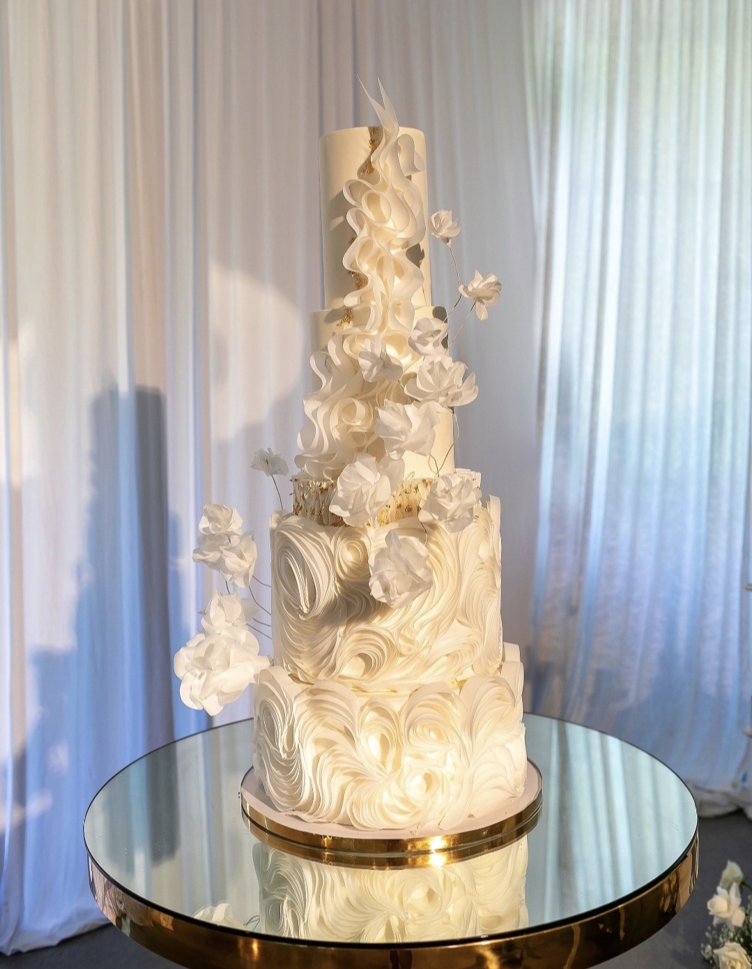 Tall, multi-tiered white wedding cake with floral decorations on a round glass table, curtains in the background.
