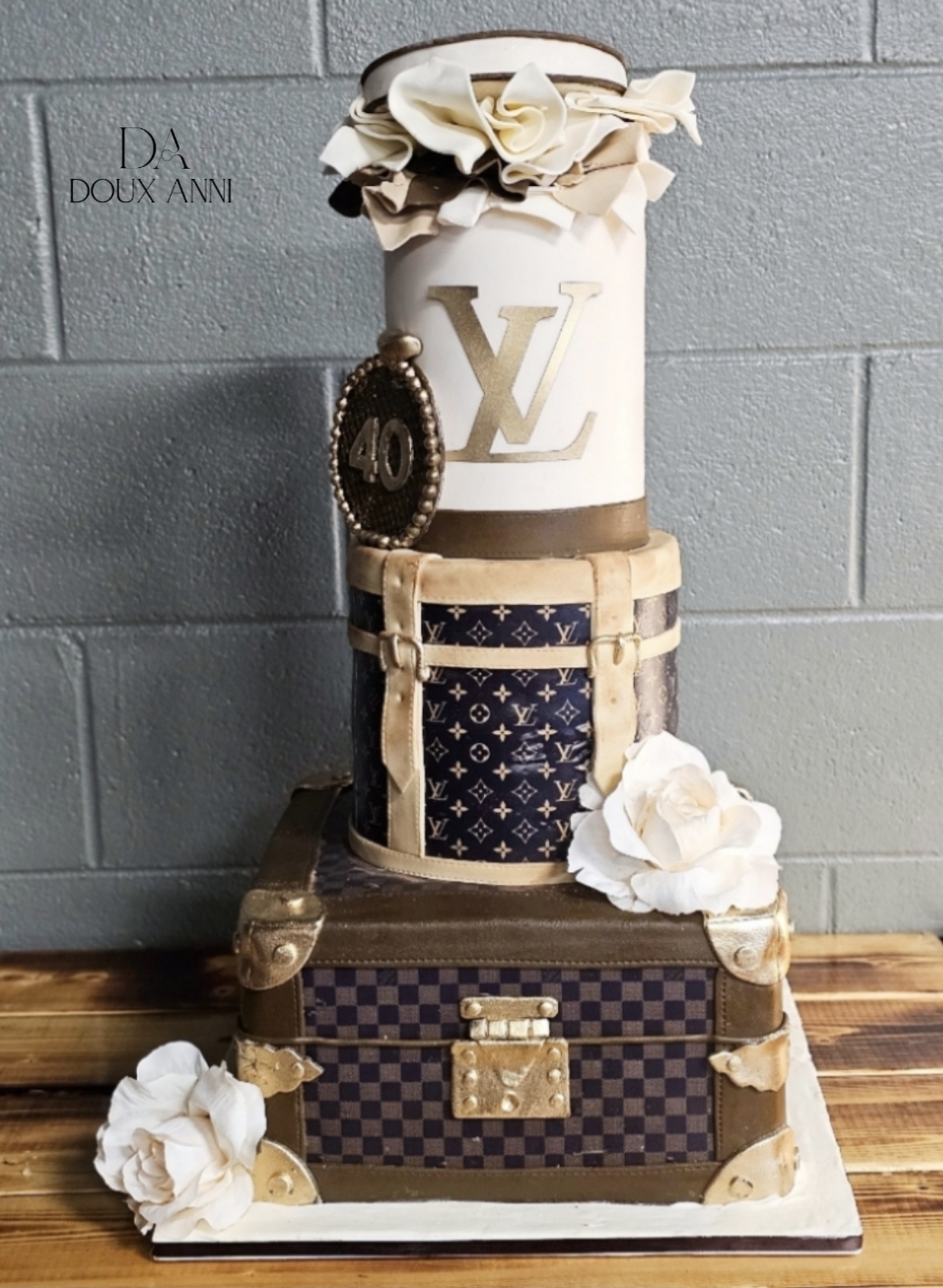 Decorative cake with Louis Vuitton patterns, flowers, and a white vase with a large flower on top, on a wooden surface.