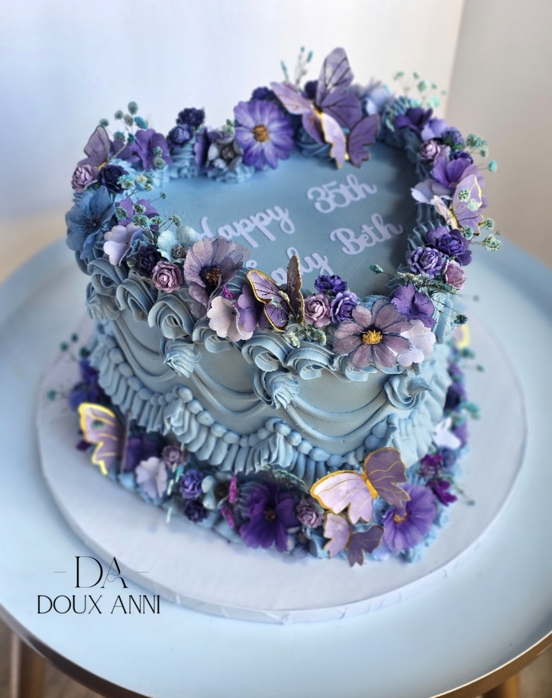 Square cake decorated with purple and blue flowers, butterflies, and a message on top, on a white cake stand.