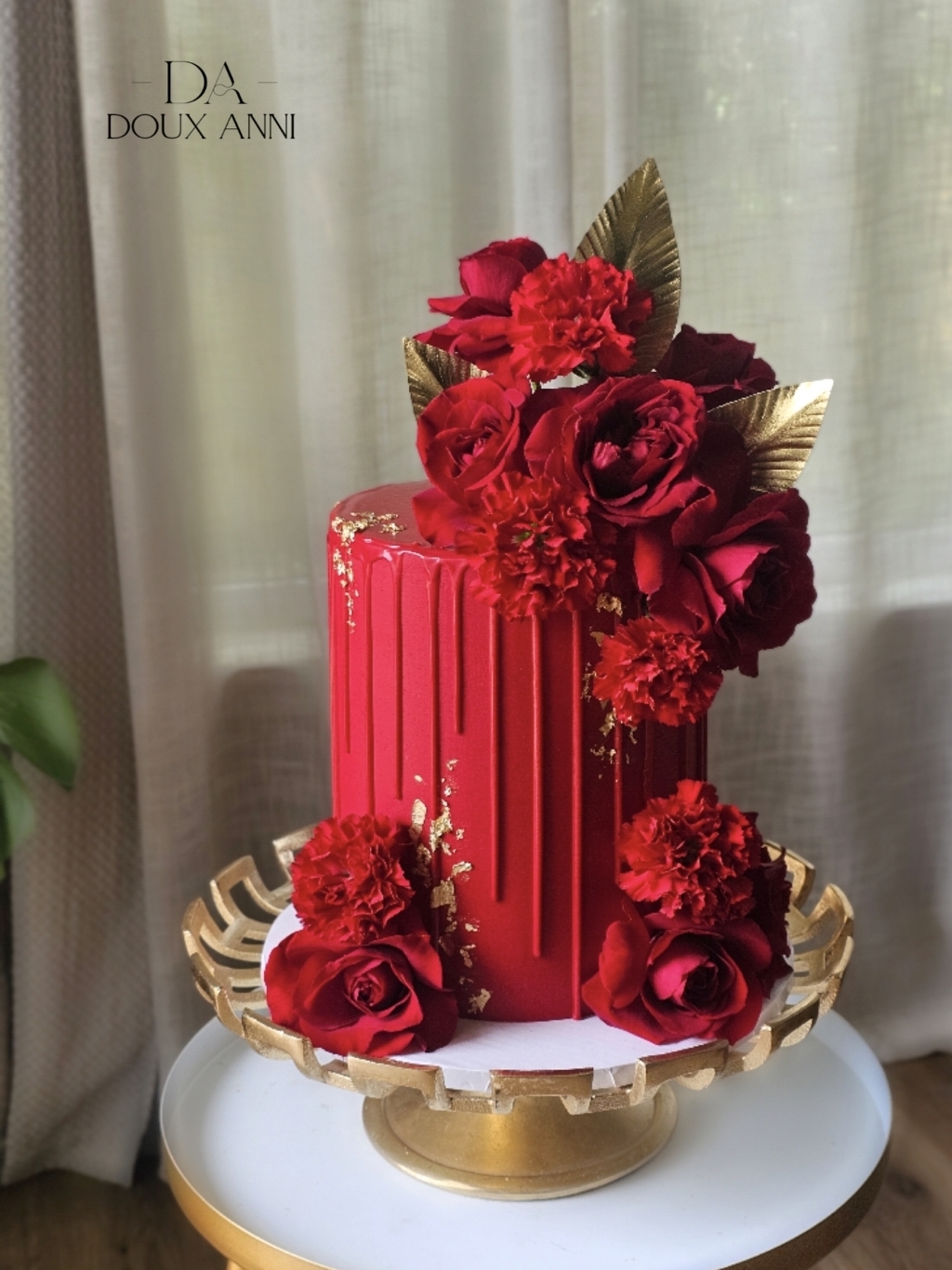 Tall red cake decorated with red flowers and gold leaves, displayed on a gold tray with a white base.