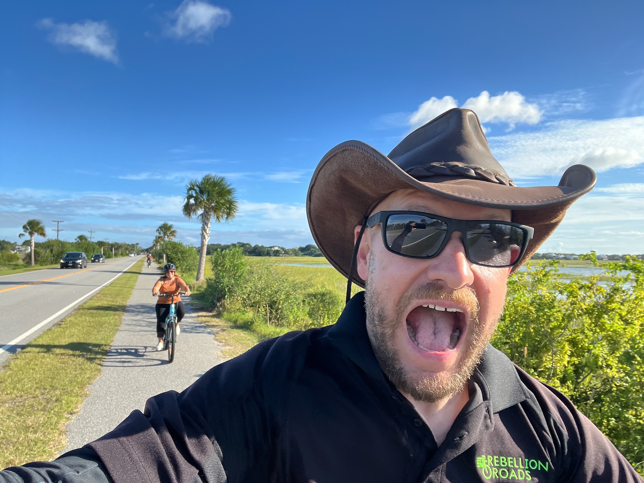 Man wearing cowboy hat and sunglasses taking a selfie outdoors with a person biking on a path behind him, blue sky, and greenery.