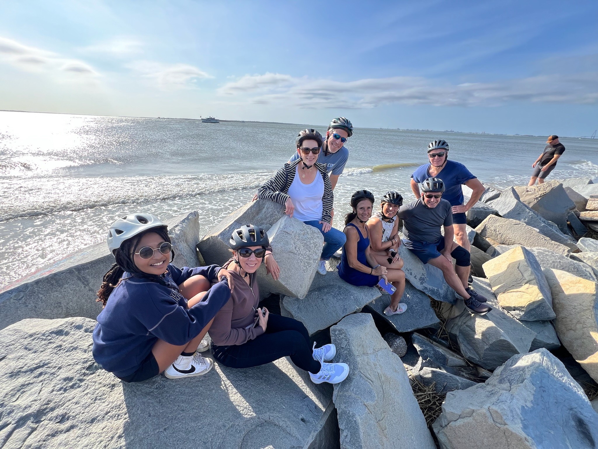 Group of seven people sitting and standing on rocks by the ocean, smiling, with a person in the background, sunny day.