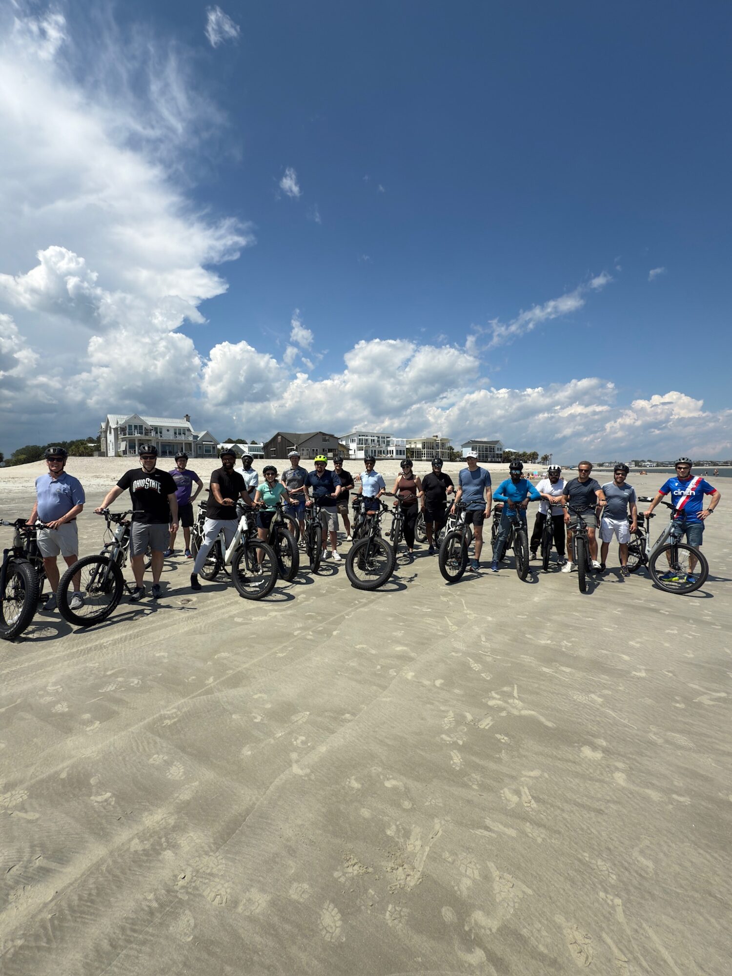 Group of people with bicycles standing on a sandy area under a blue sky with clouds.