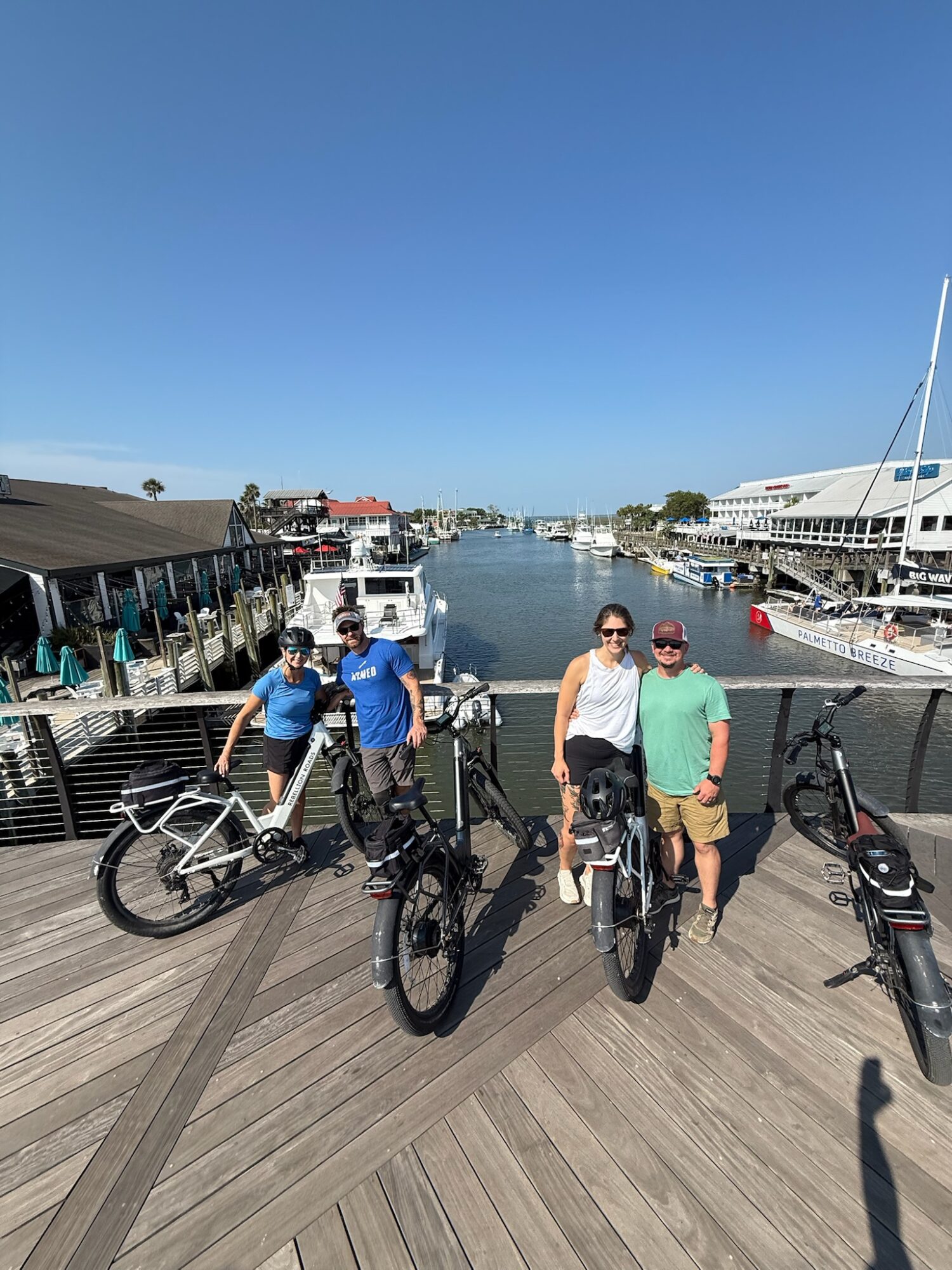 Three people with bicycles on a dock by a marina, with boats and buildings in the background under a clear blue sky.