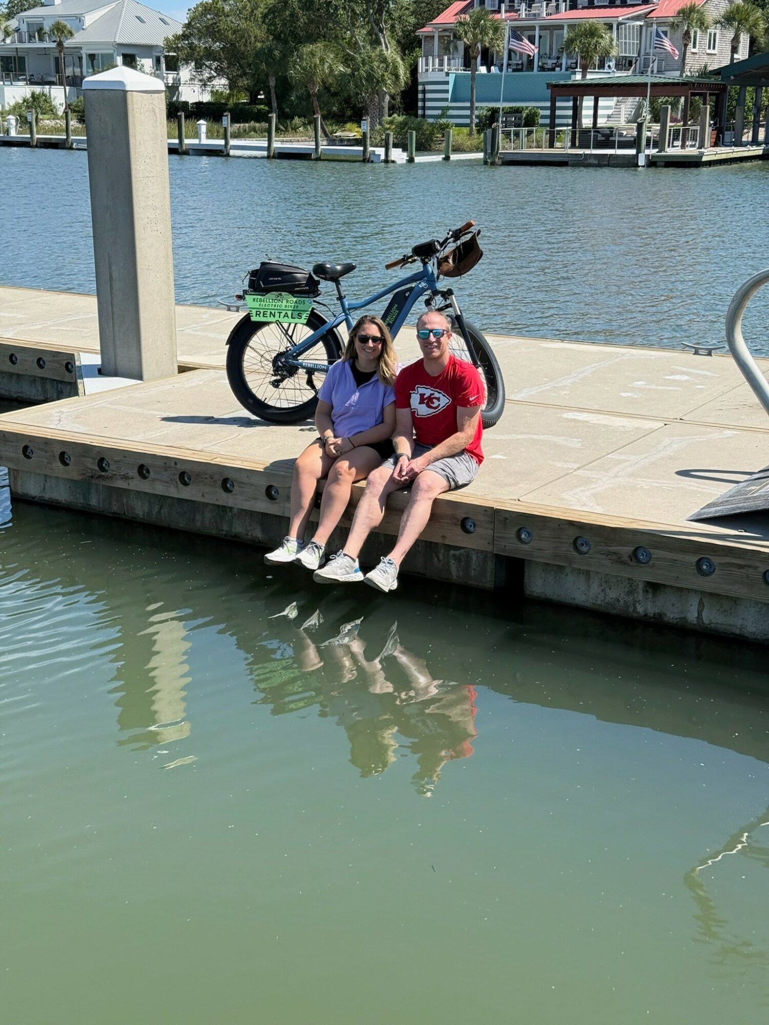 Two people sit on a dock by water, with a bicycle behind them and houses in the background.