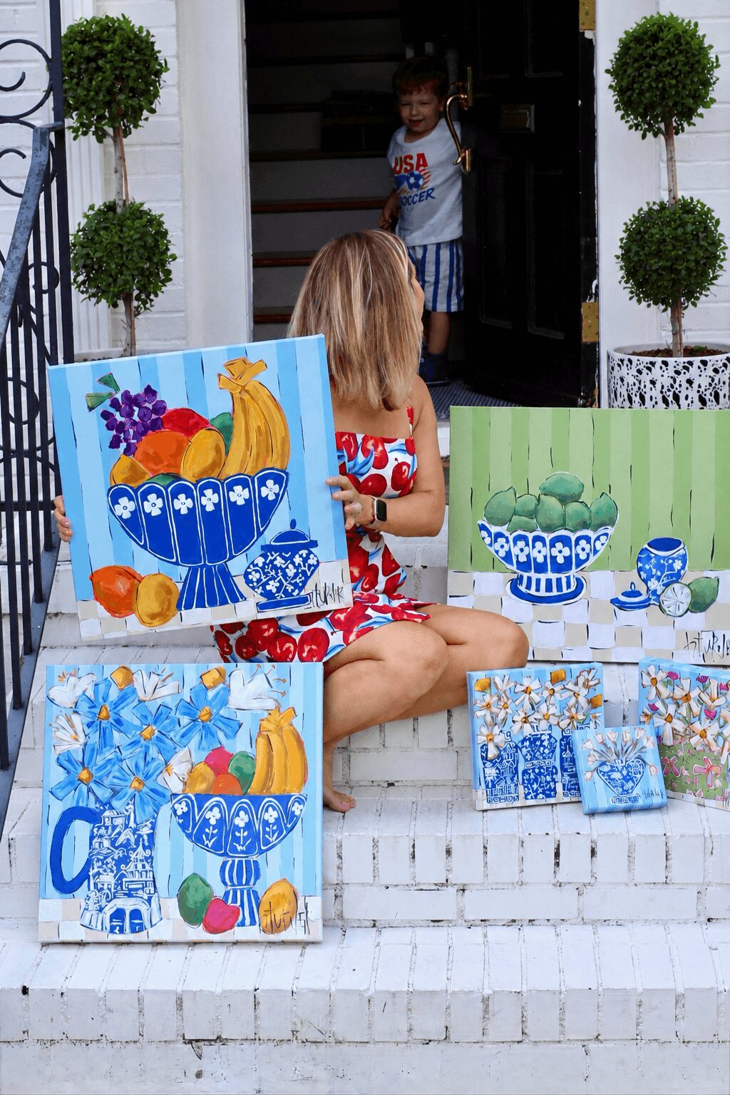 Child with blonde hair sitting on steps, surrounded by colorful paintings of fruit and flowers, with a girl standing in doorway behind.