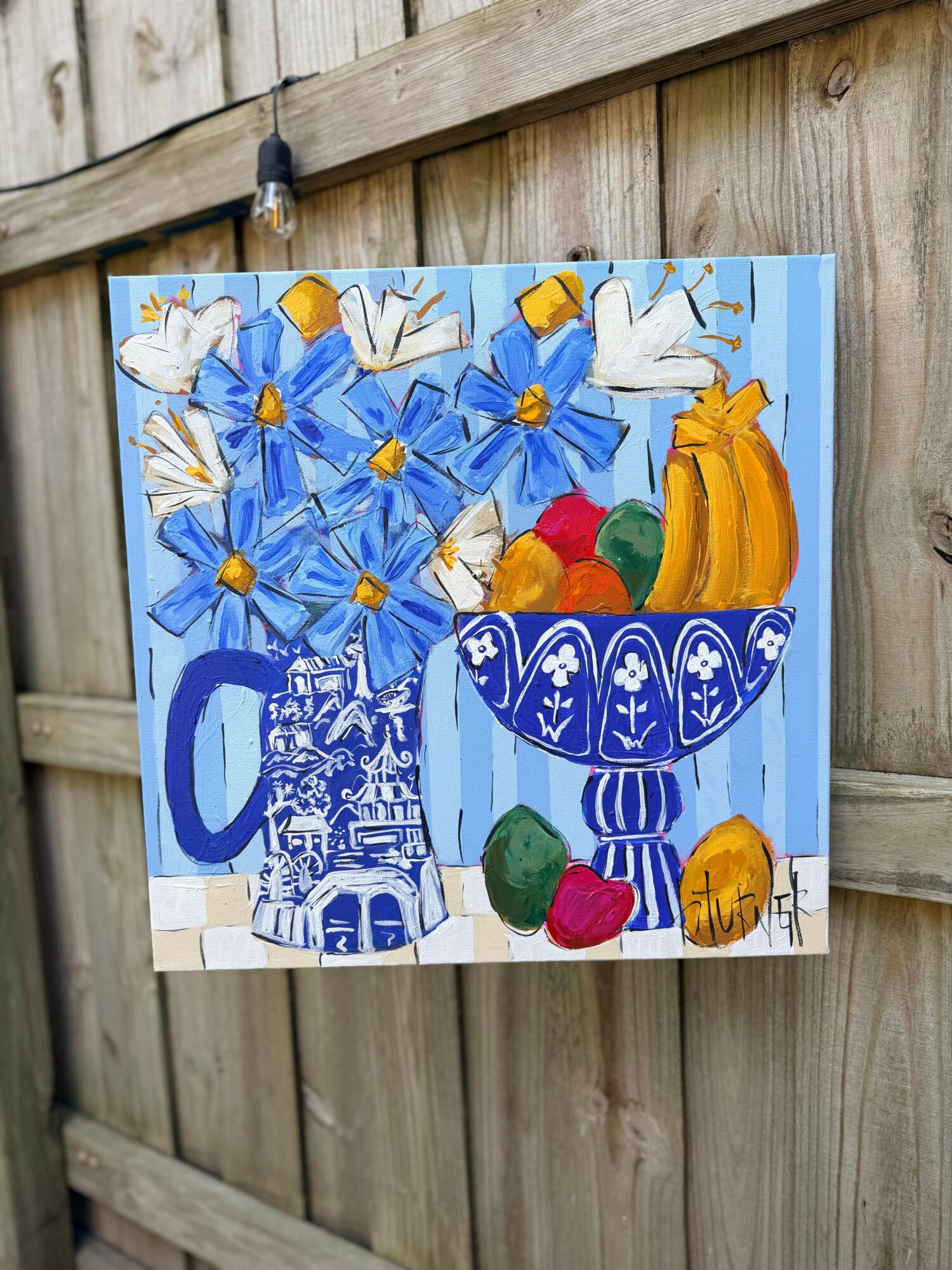 Painting of blue flowers, a bowl of fruit, and a blue patterned pitcher on a wooden fence.
