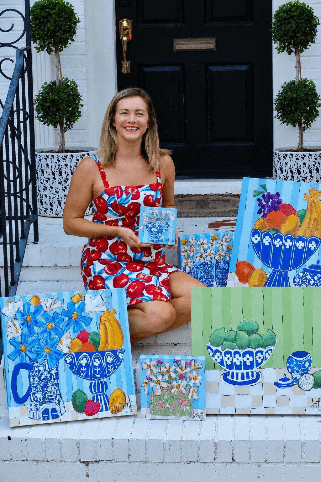 Woman sitting on steps with colorful painted pots and artwork, smiling, in front of a black door with potted plants.
