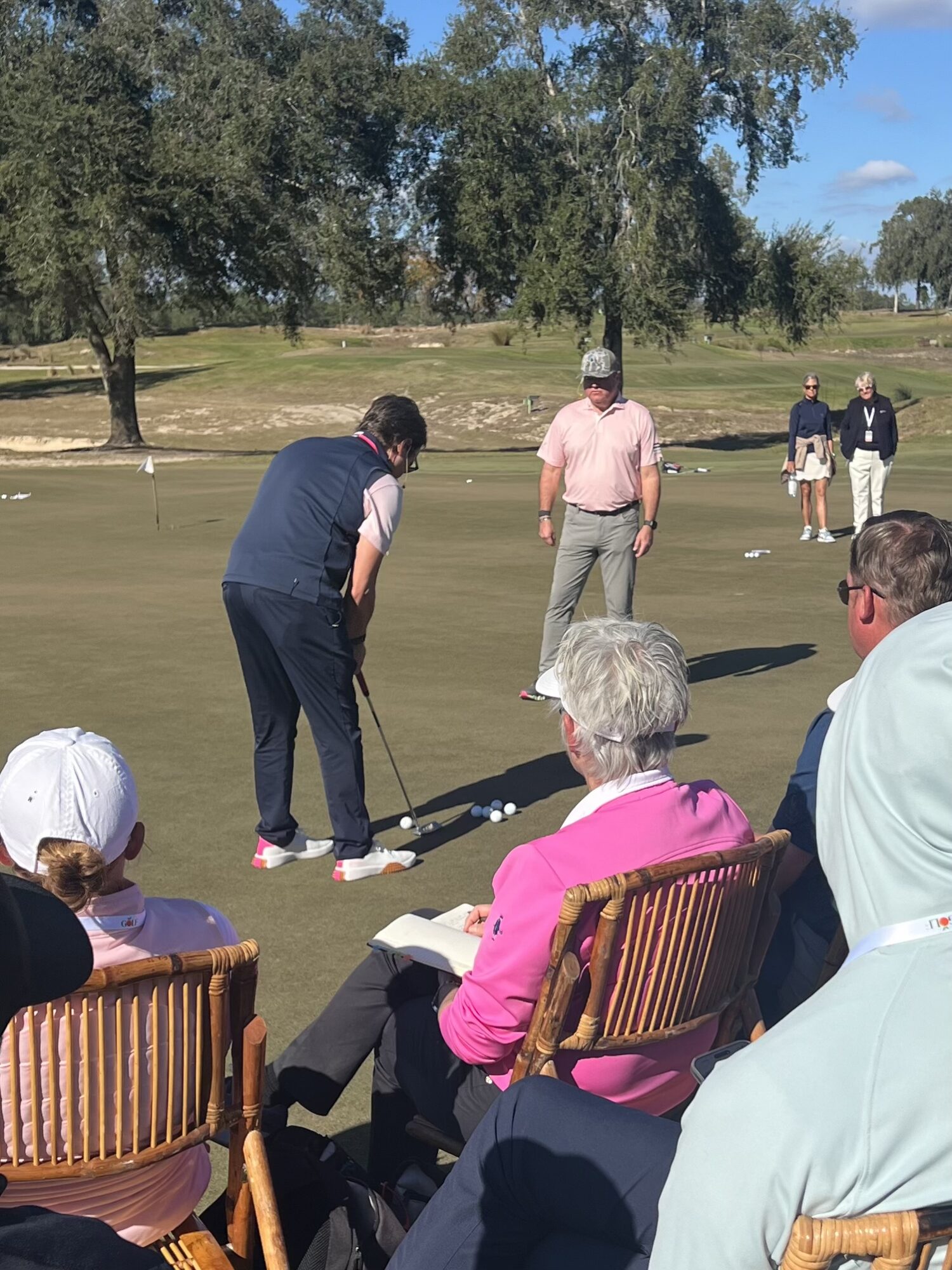 People on a golf course preparing to putt, with trees and a blue sky in the background.