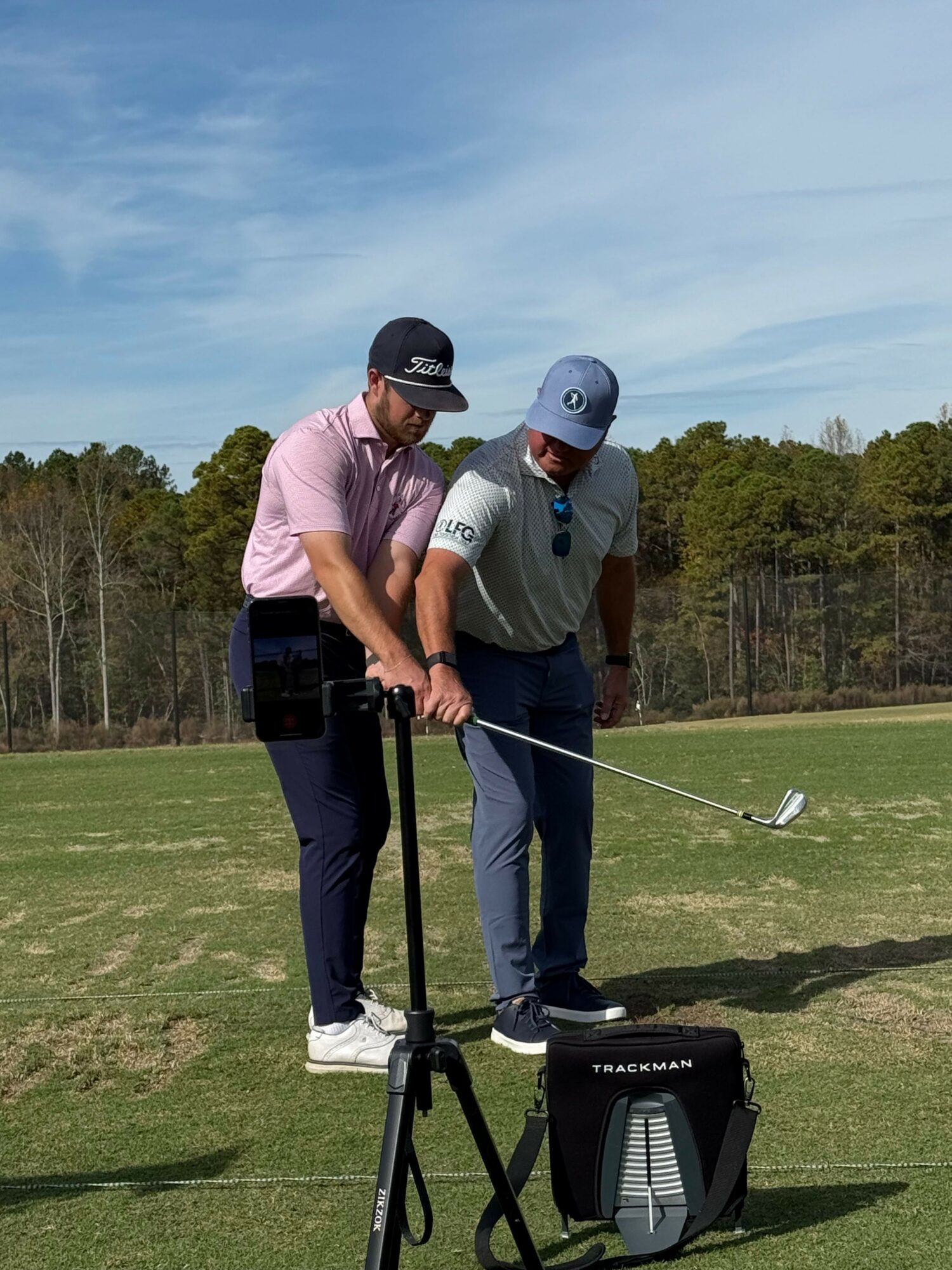 Two people playing golf on a course, one preparing to swing, with trees and blue sky in background.