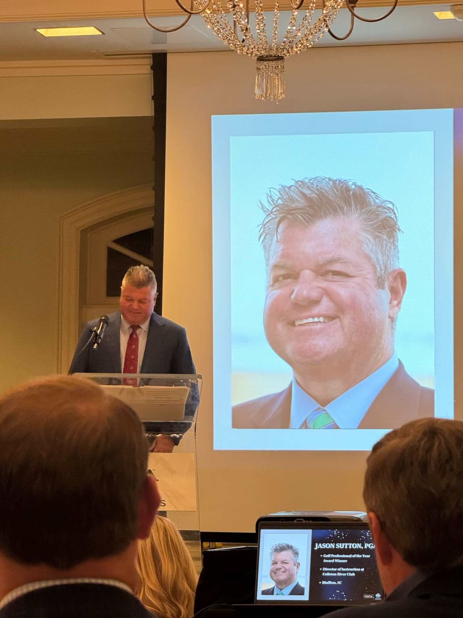 Person giving a speech at a podium in front of a large screen displaying a smiling man's face, audience members visible.