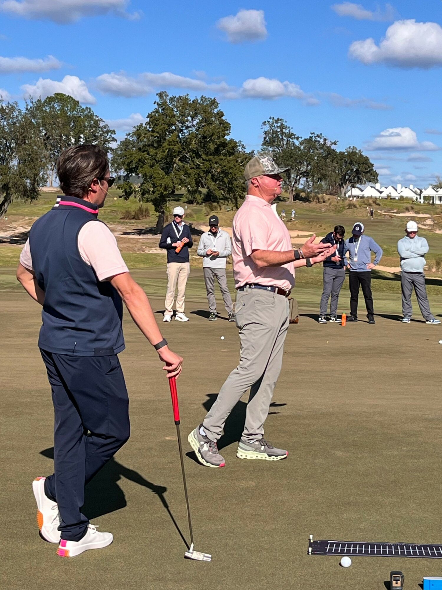 Person preparing to putt on a golf course with several people watching, trees, and blue sky in background.