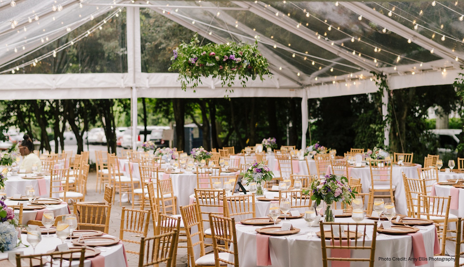 Elegant event setup with round tables, chairs, floral centerpieces, and hanging lights inside a glass tent.
