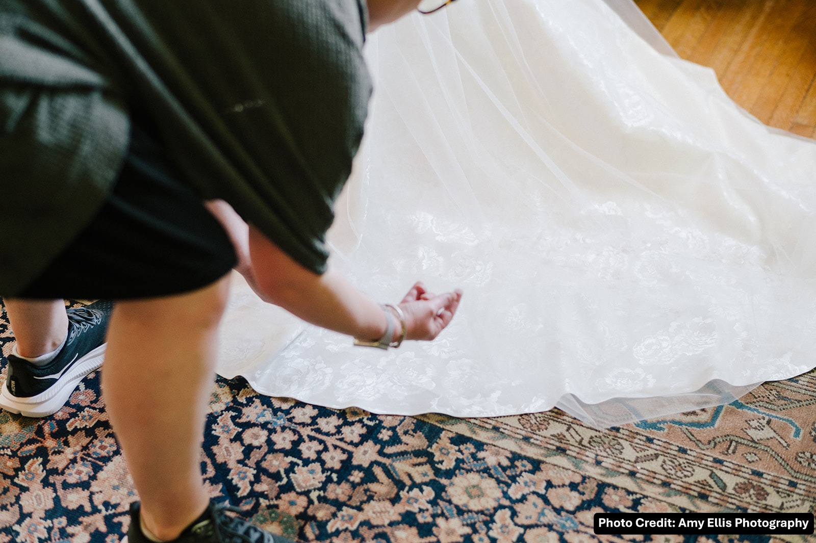 Person bending over on a patterned rug, reaching toward a white fabric or dress on a wooden floor.