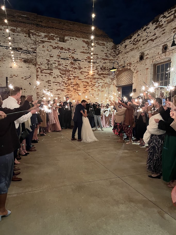People dancing and holding sparklers in a rustic indoor venue with brick walls and string lights.