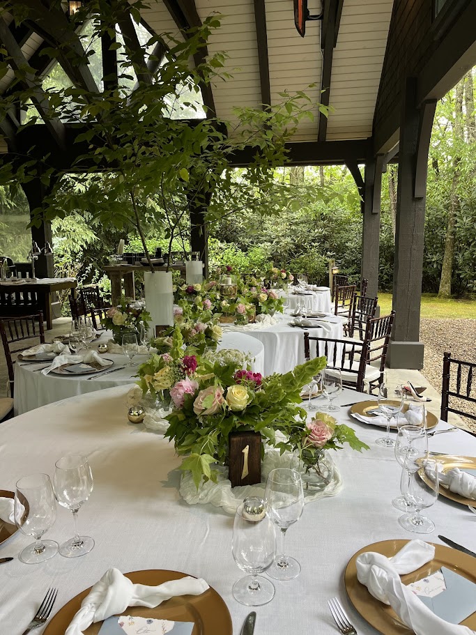 Elegant outdoor dining setup with floral centerpiece, white tablecloths, and chairs under a covered patio, surrounded by greenery.