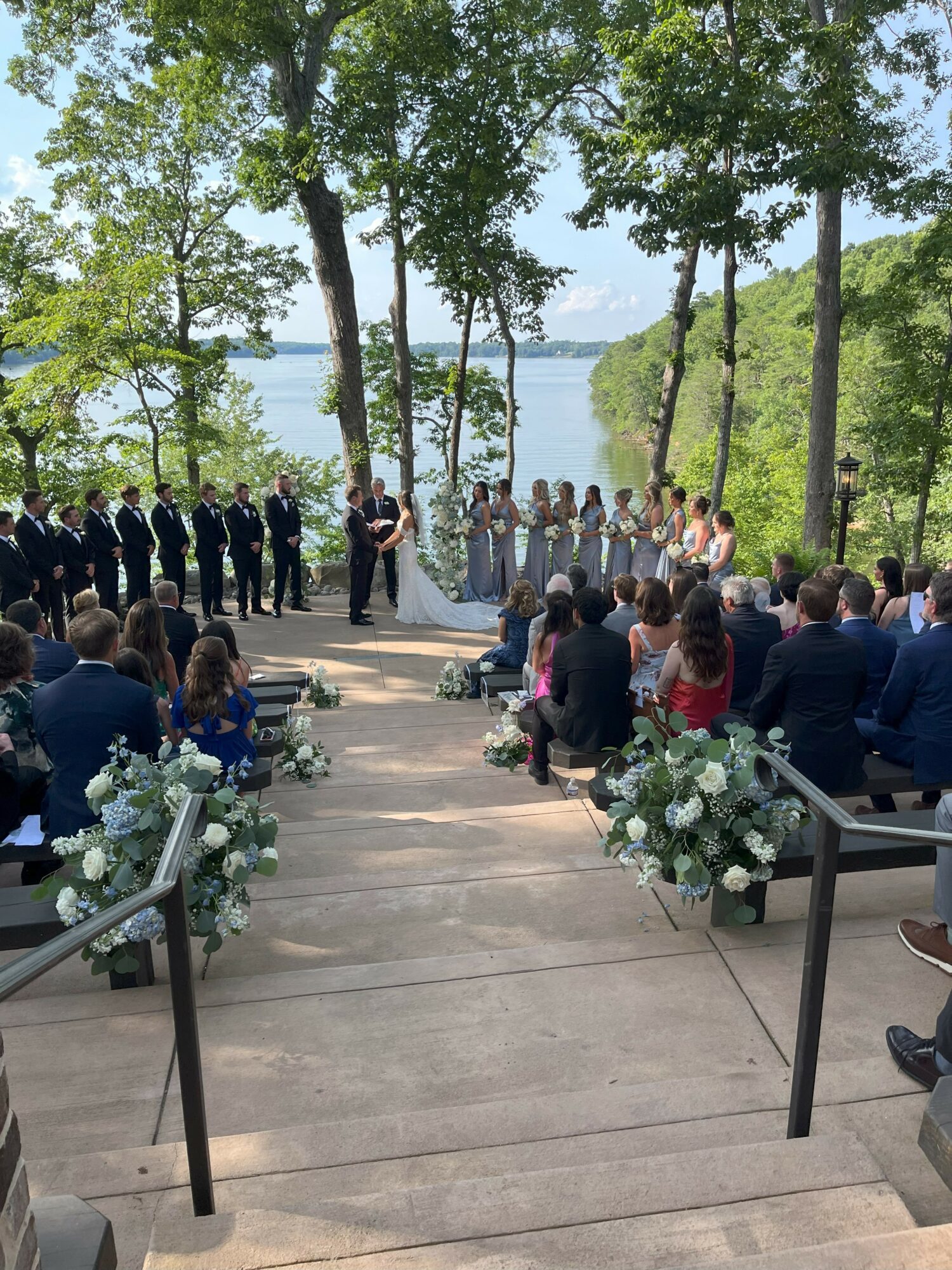 Outdoor wedding ceremony with guests seated, officiant, and wedding party near a lake surrounded by trees.