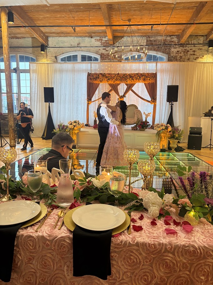 People dancing on stage with a decorated backdrop, floral arrangements on a table in foreground, in a rustic venue.