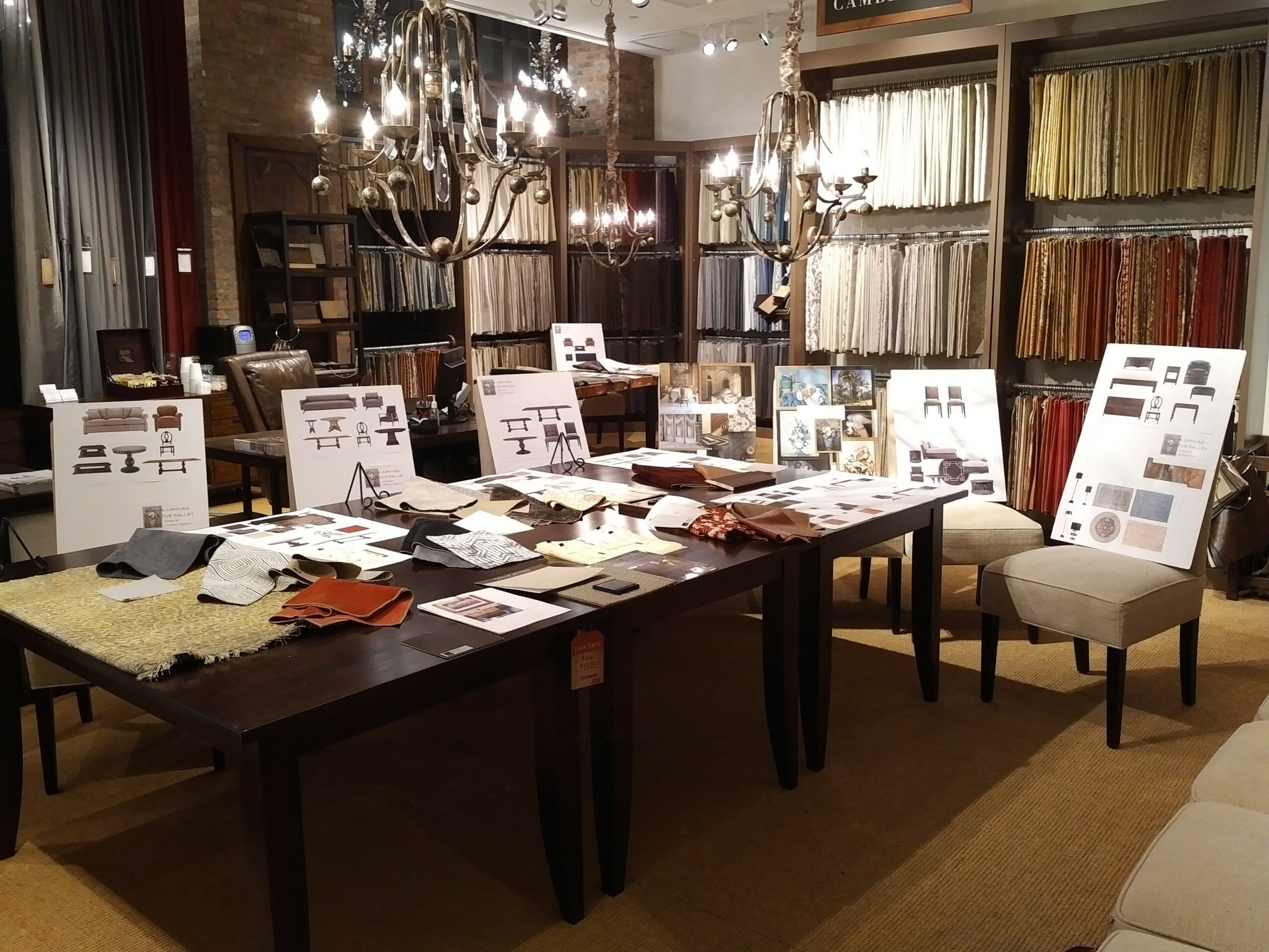 Interior of a fabric or wallpaper store with tables, chairs, and fabric samples displayed, chandeliers hanging from the ceiling.