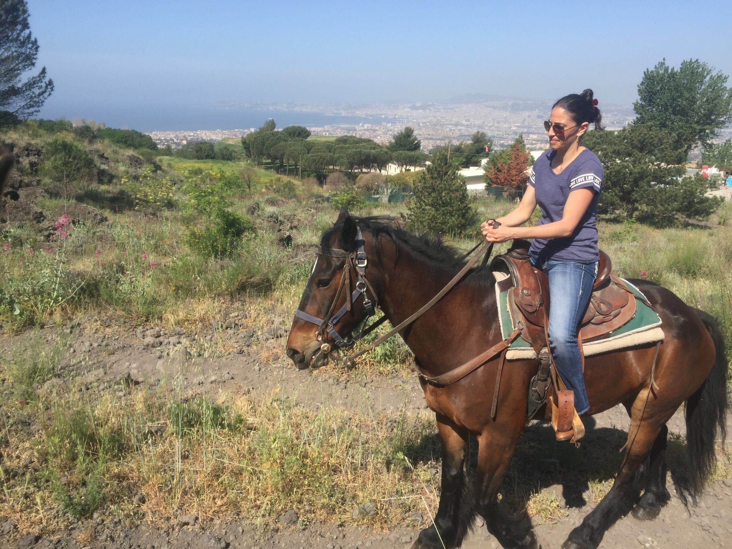 Person riding a horse outdoors with a landscape background, wearing sunglasses and casual clothing.