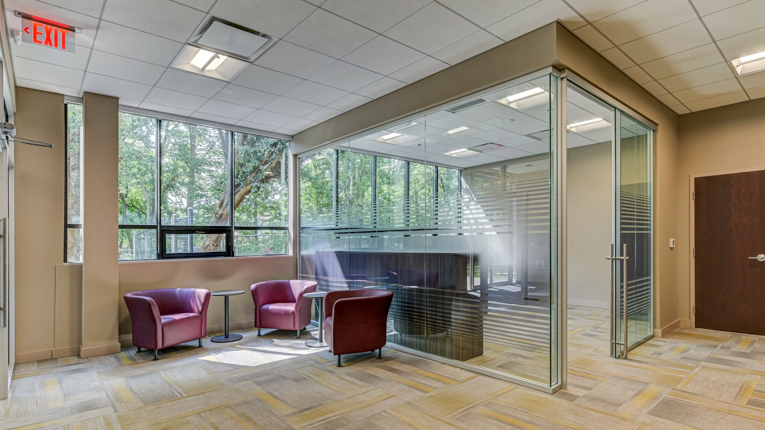 Lobby area with two purple chairs and a glass-walled office space, large windows showing trees outside, and a wooden door.