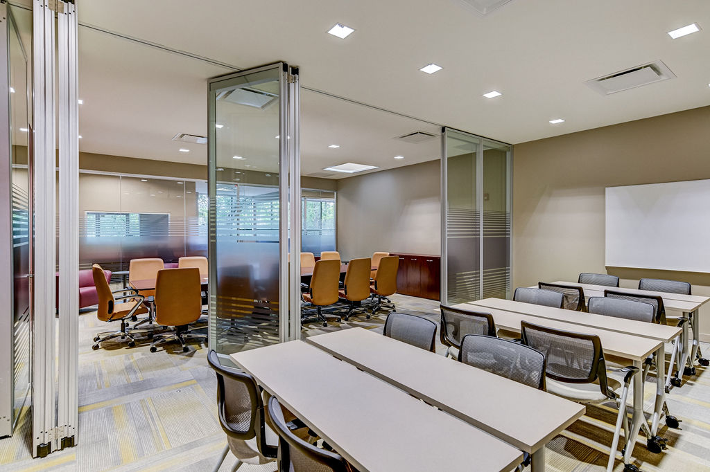 Conference room with tables, chairs, glass partitions, and a whiteboard, illuminated by ceiling lights.