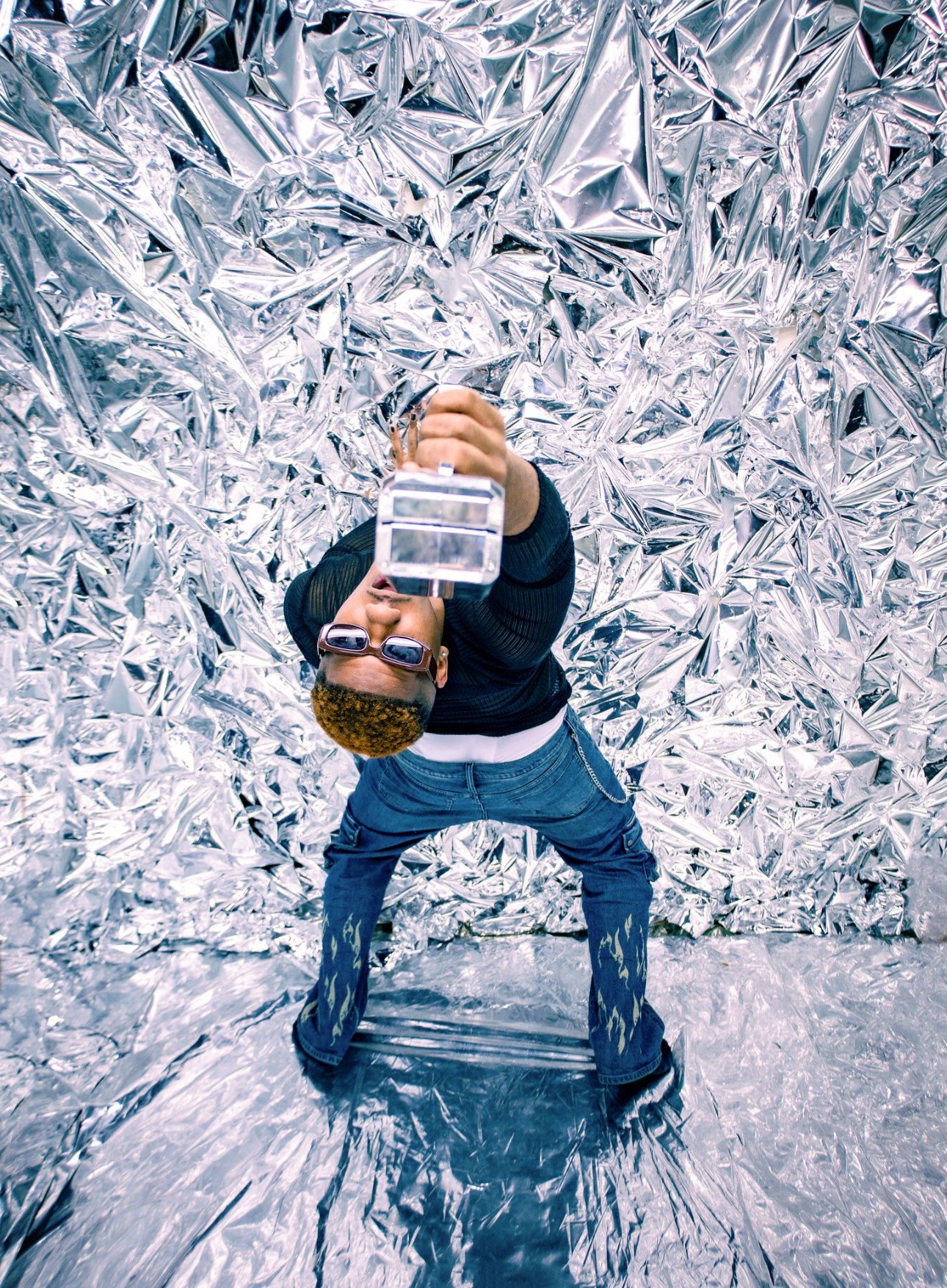 Person taking a photo with a camera, standing in front of crinkled metallic background, legs apart, head tilted downward.