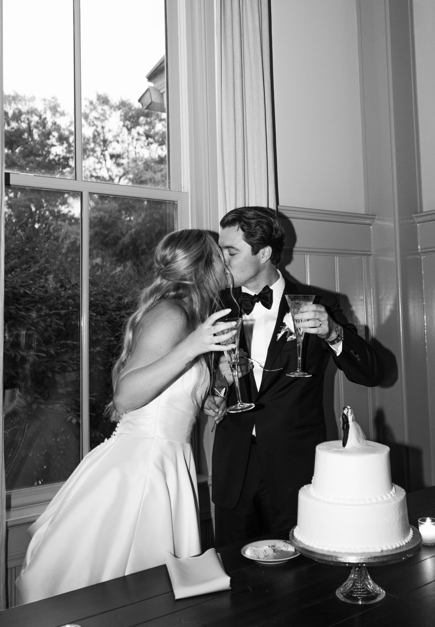 A couple in formal attire sharing a kiss at a celebration with a cake on the table.