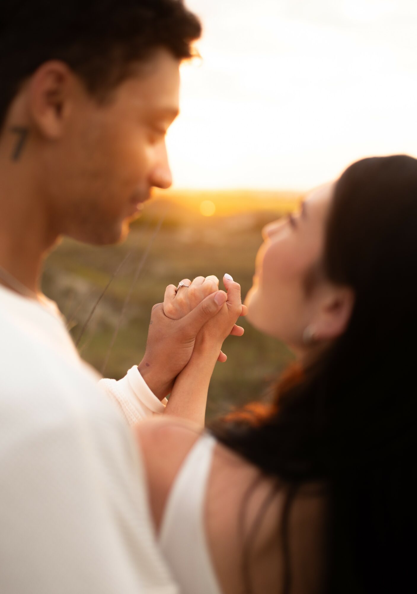 A man and woman hold hands close, facing each other outdoors during sunset, with blurred landscape background.